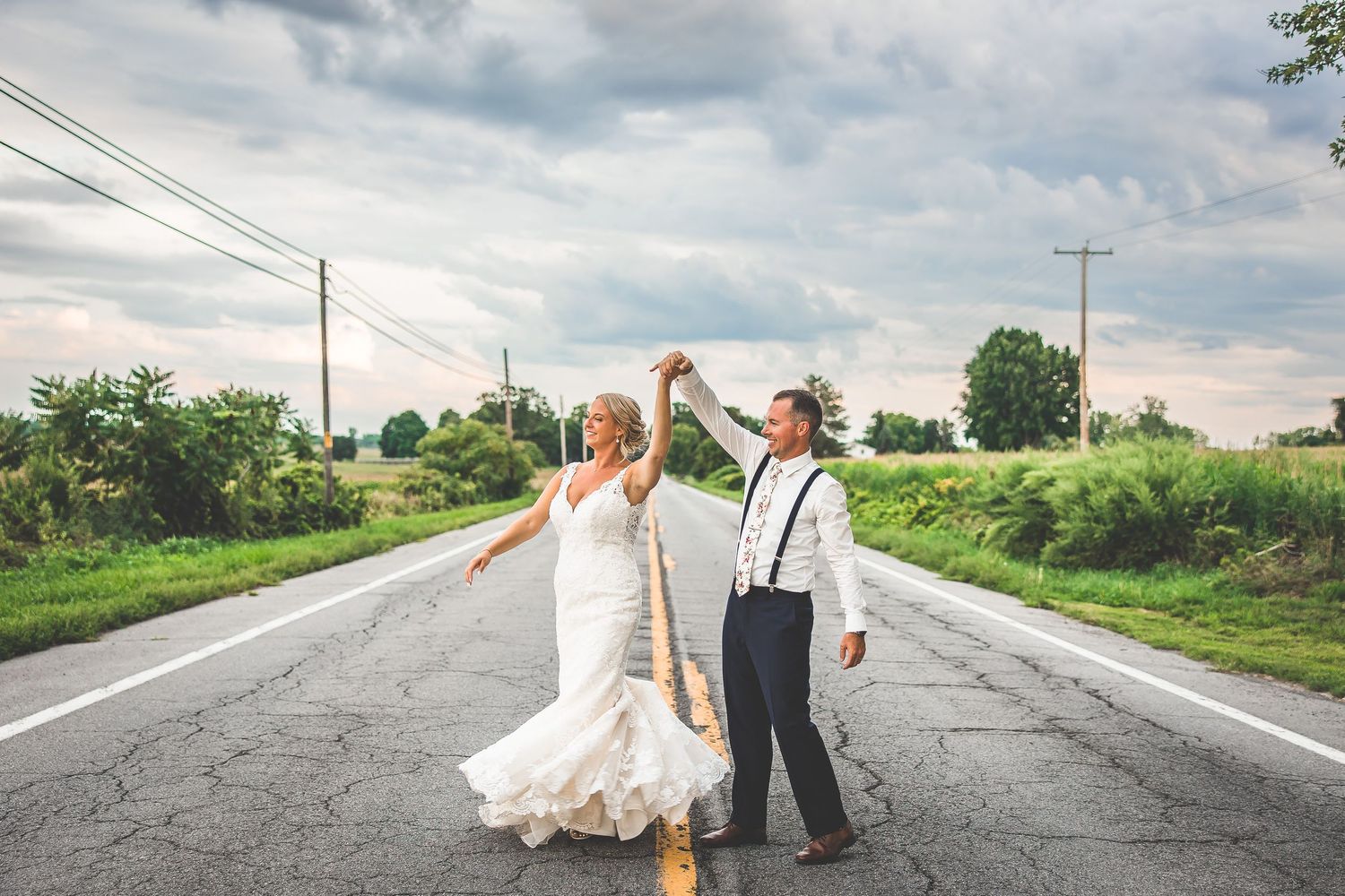 Wedding couple dancing on a rural road surrounded by green fields.