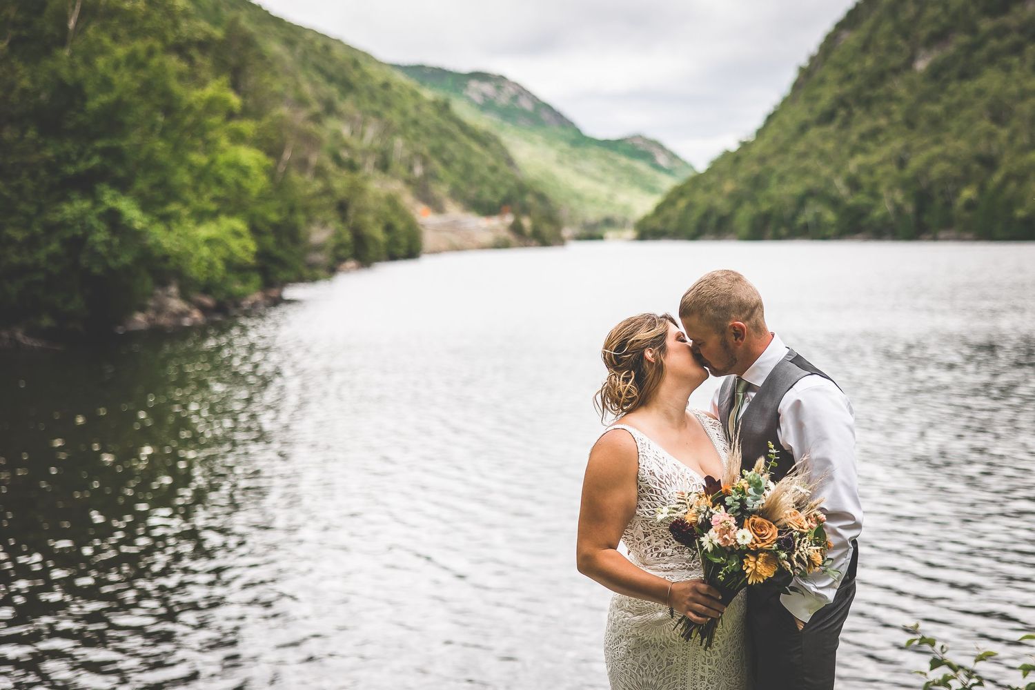 Wedding couple shares intimate moment by mountain lake under cloudy skies.