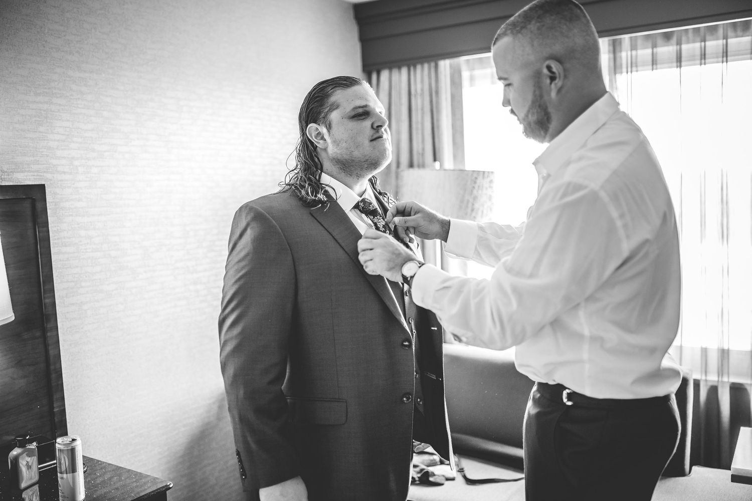 Two people adjusting formal wear in a hotel room with natural window light.
