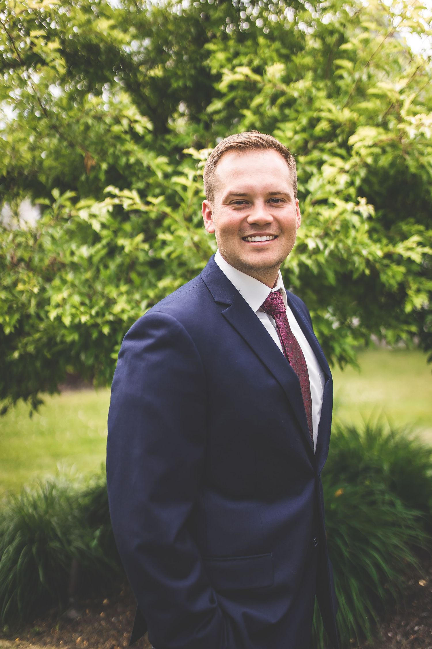 Wedding party member in navy suit and burgundy tie posing in garden setting.