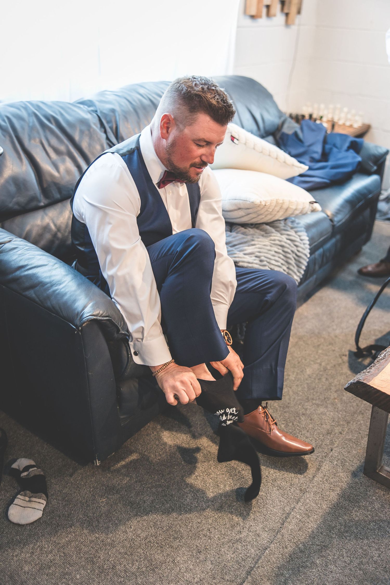 Groom putting on dress shoes while sitting on leather couch during wedding preparations.