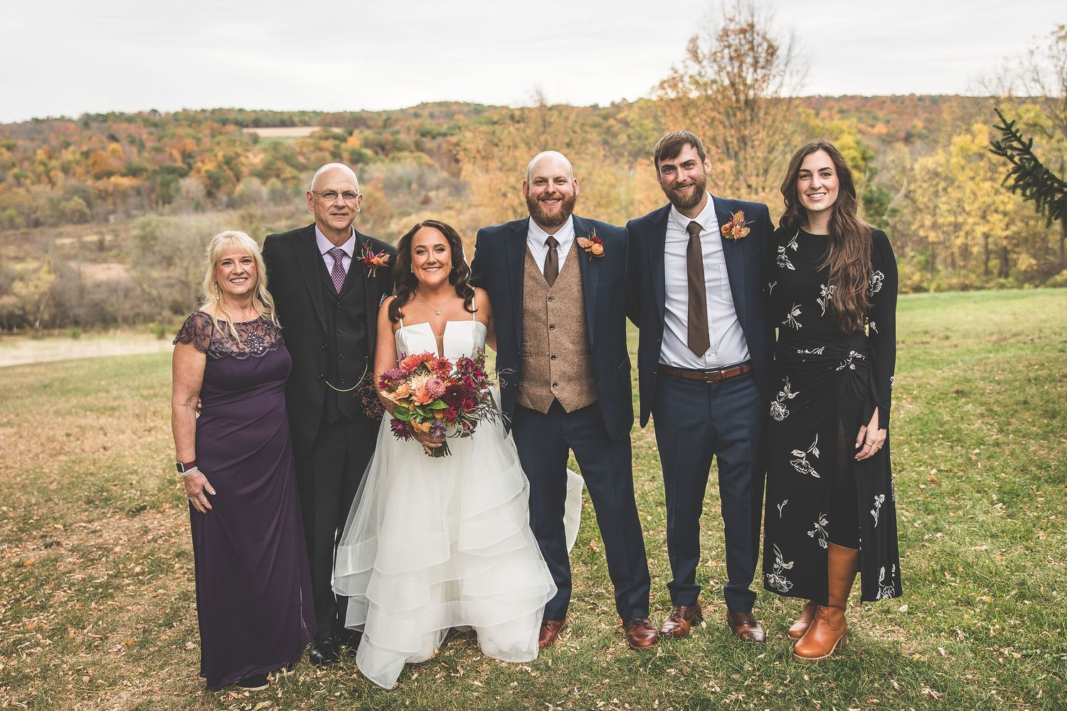 Wedding party members pose together in autumn field with fall foliage in background.