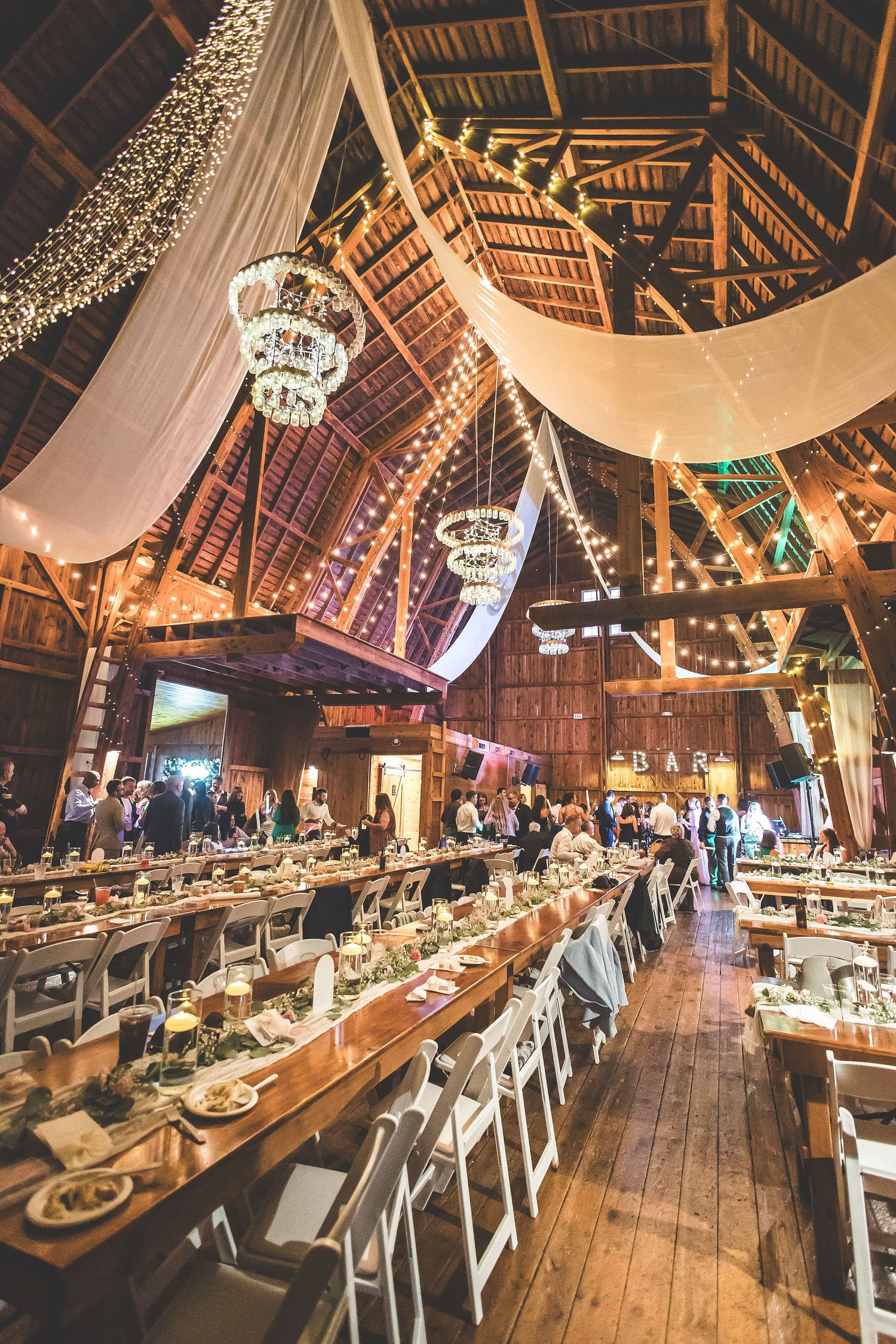 Elegant barn wedding reception setup with long tables, white linens, and twinkling lights overhead.