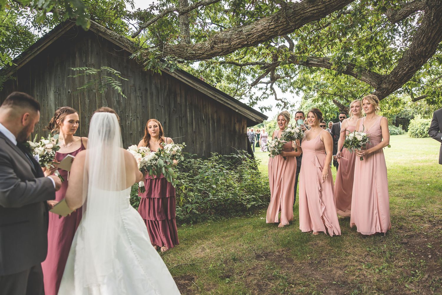 Wedding party in pink dresses and grey suits gathered outdoors near a rustic barn.