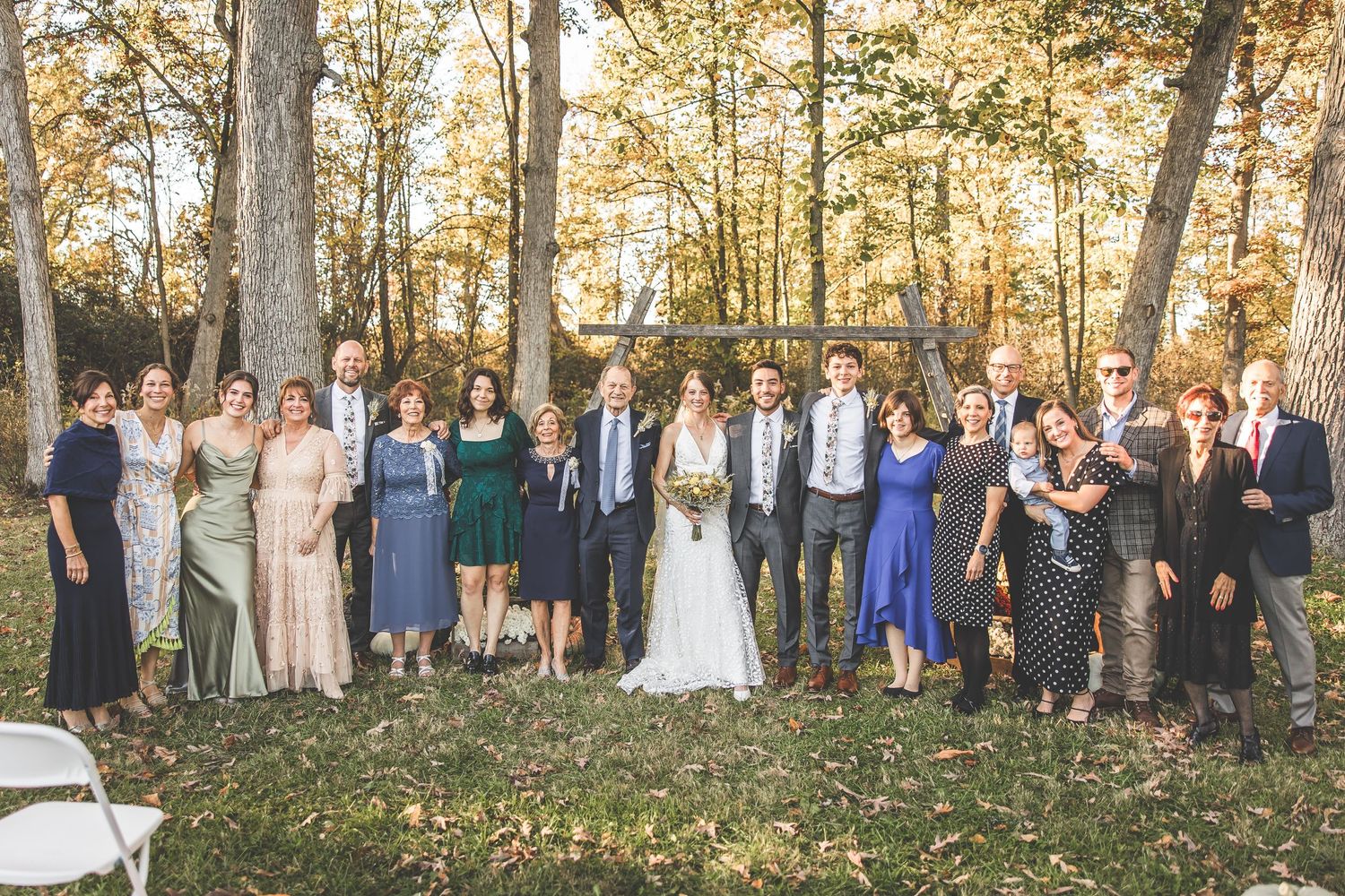 Extended family and wedding guests pose for a large group photo in a wooded autumn setting.