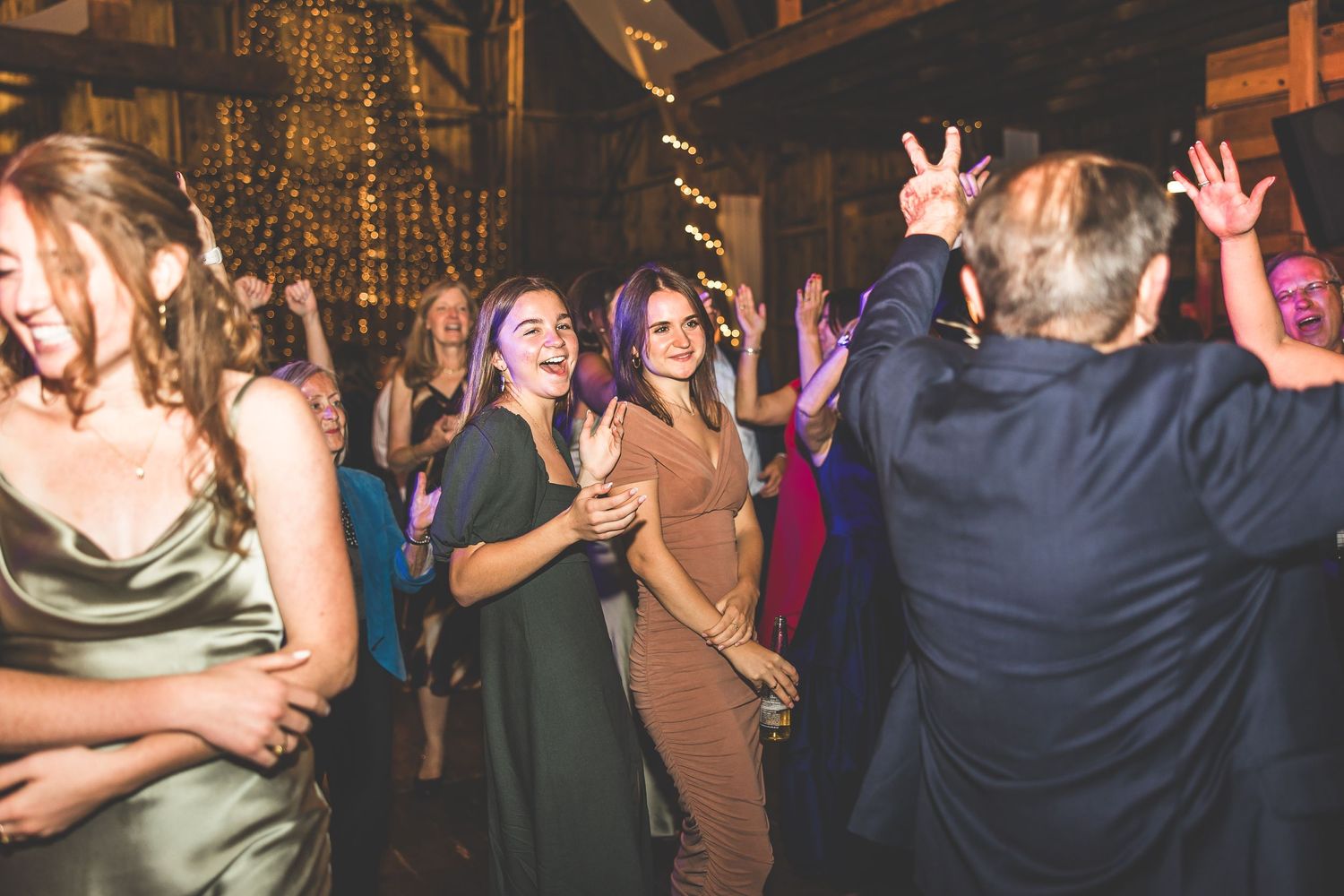 Energetic crowd dancing and celebrating at a wedding reception in a rustic barn setting.
