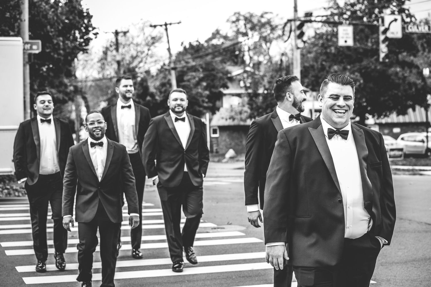 Groomsmen in formal black tuxedos walking across a city street.