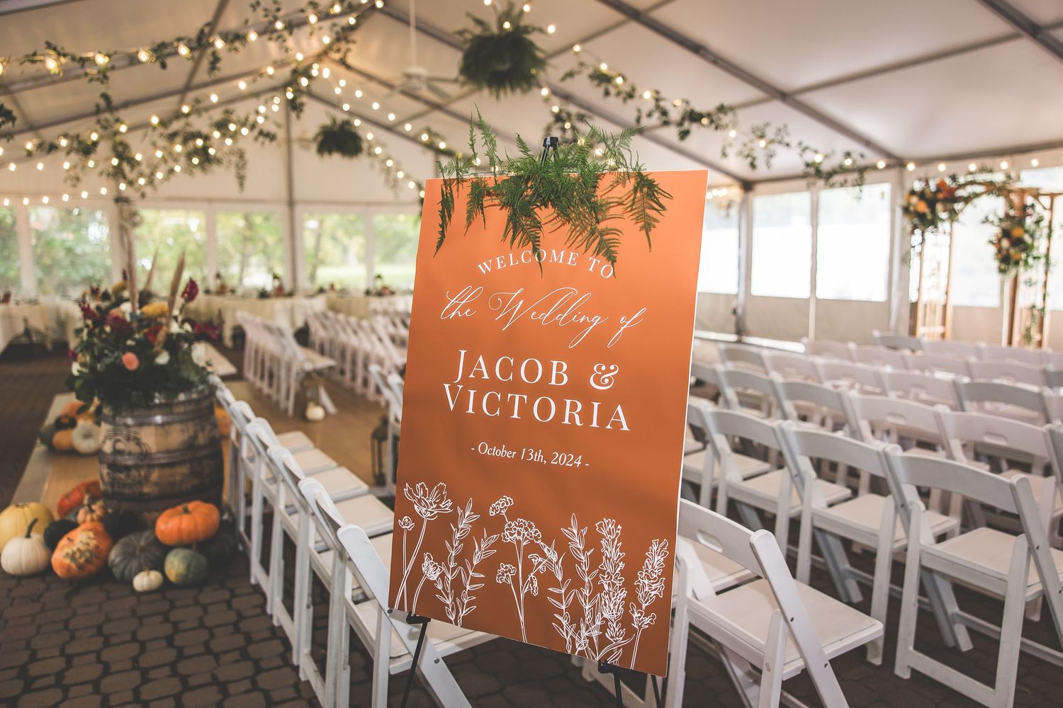 Rustic wedding ceremony setup with white chairs and terracotta welcome sign in a tented venue.