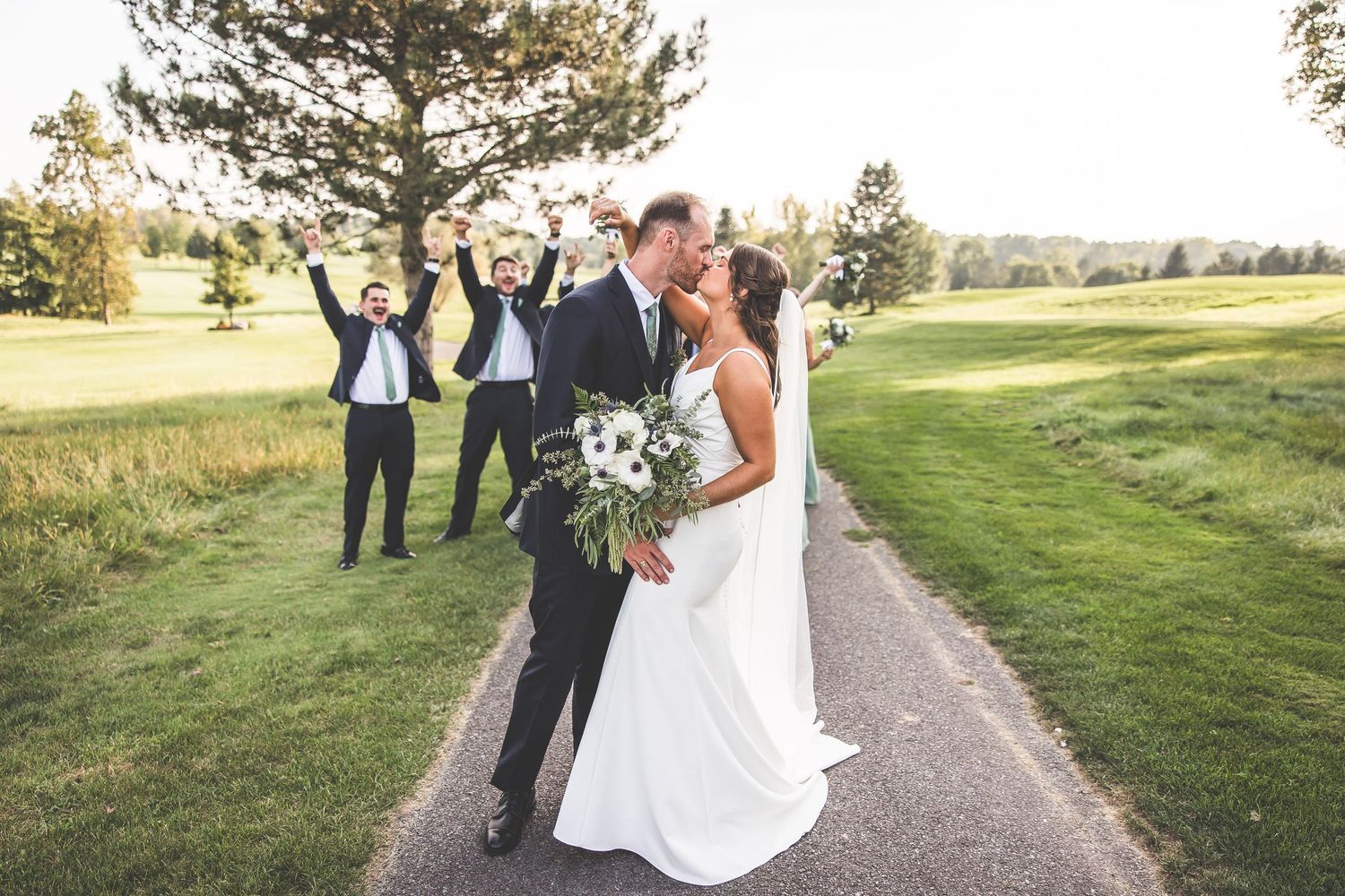 Newlyweds share romantic moment on path while wedding party celebrates in background.