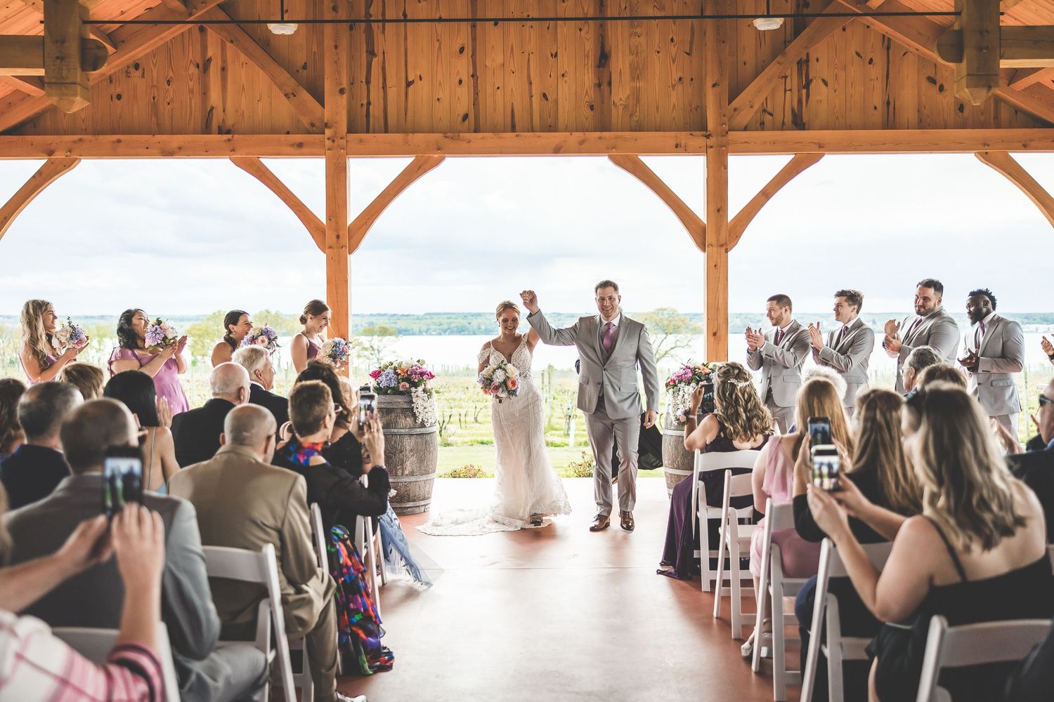 Wedding ceremony in progress inside rustic wooden pavilion with guests seated on white chairs.