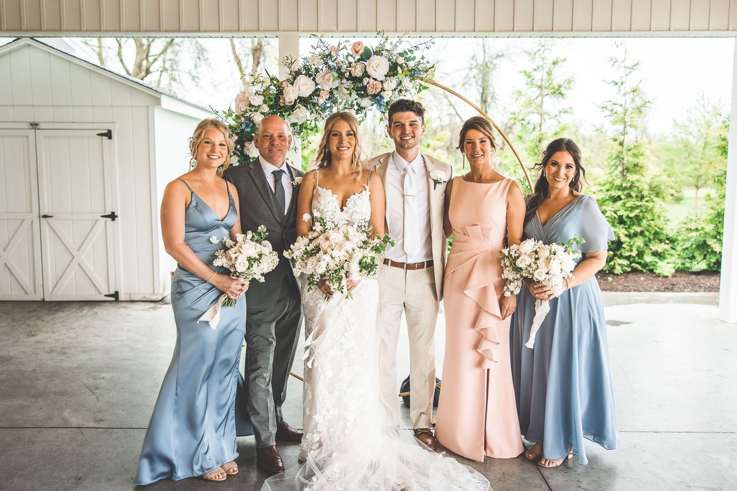 Wedding party poses in formal attire with bridesmaids in blue and pink dresses under a covered outdoor area.