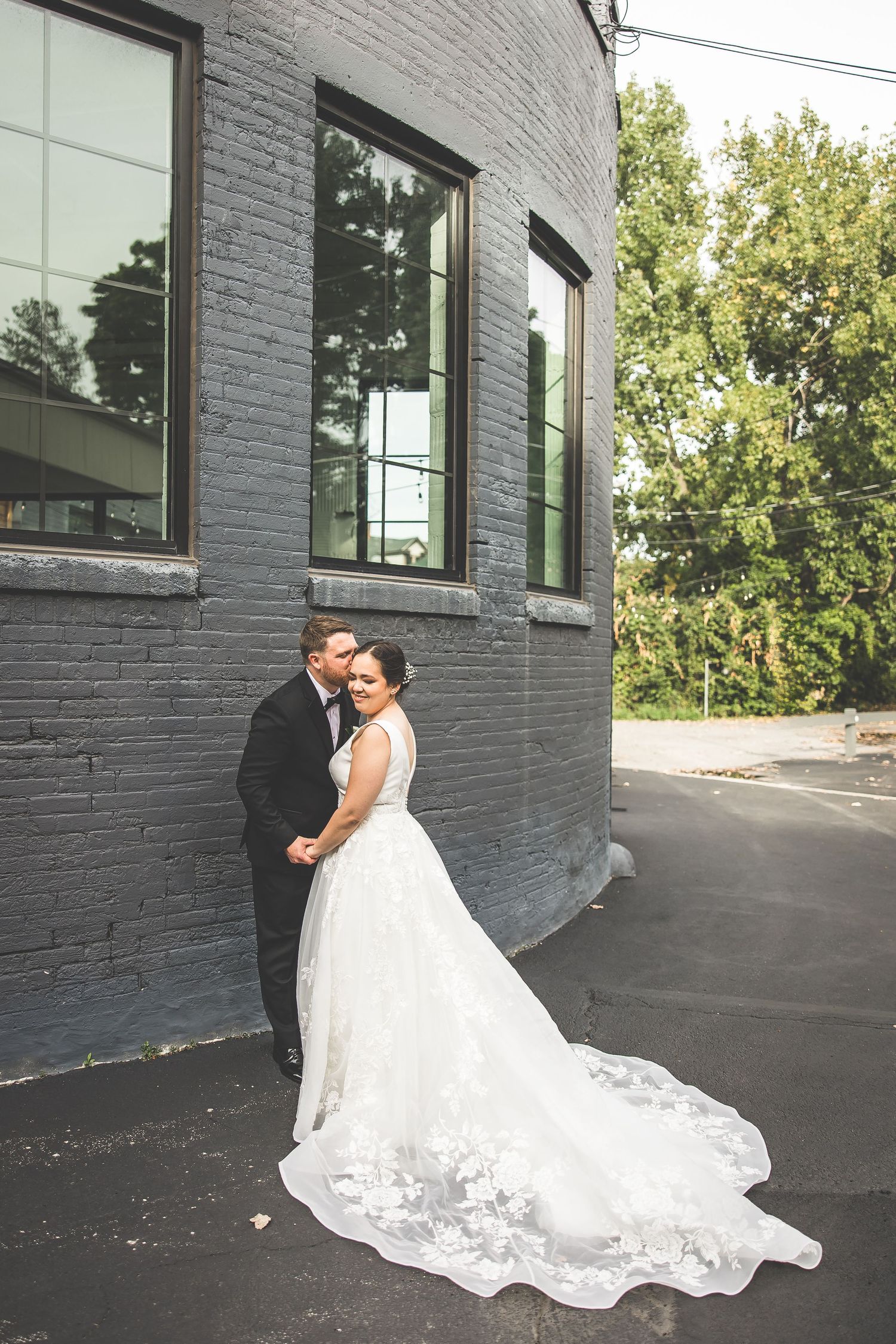 Elegant wedding dress with lace train flowing across dark pavement against grey brick wall.