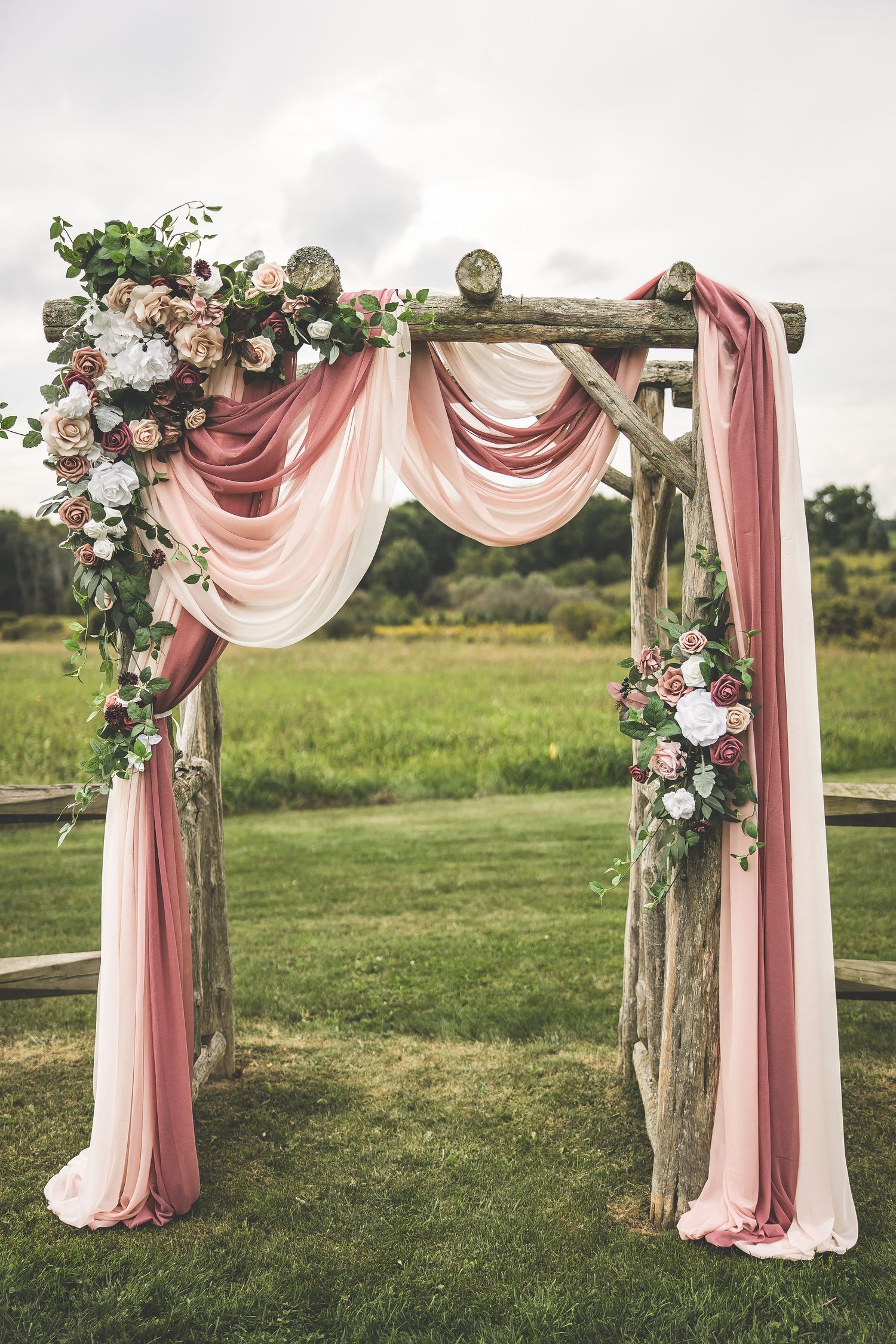 Rustic wedding arch decorated with pink draping fabric and floral arrangements in an outdoor setting.