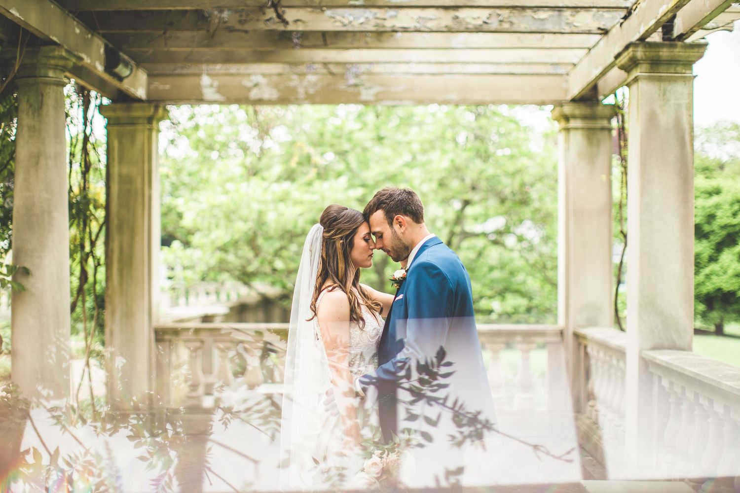 A romantic wedding moment captured under a columned pergola surrounded by lush greenery.
