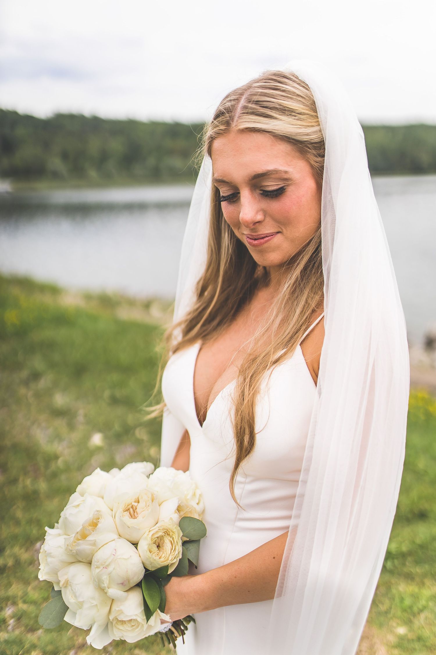 Elegant bridal portrait featuring a white wedding dress and bouquet of cream roses against a lakeside setting.
