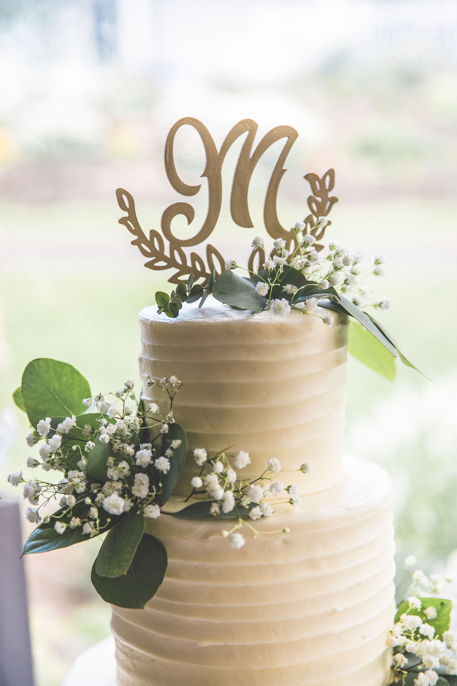 White wedding cake decorated with baby's breath, eucalyptus leaves and a gold monogram M topper.
