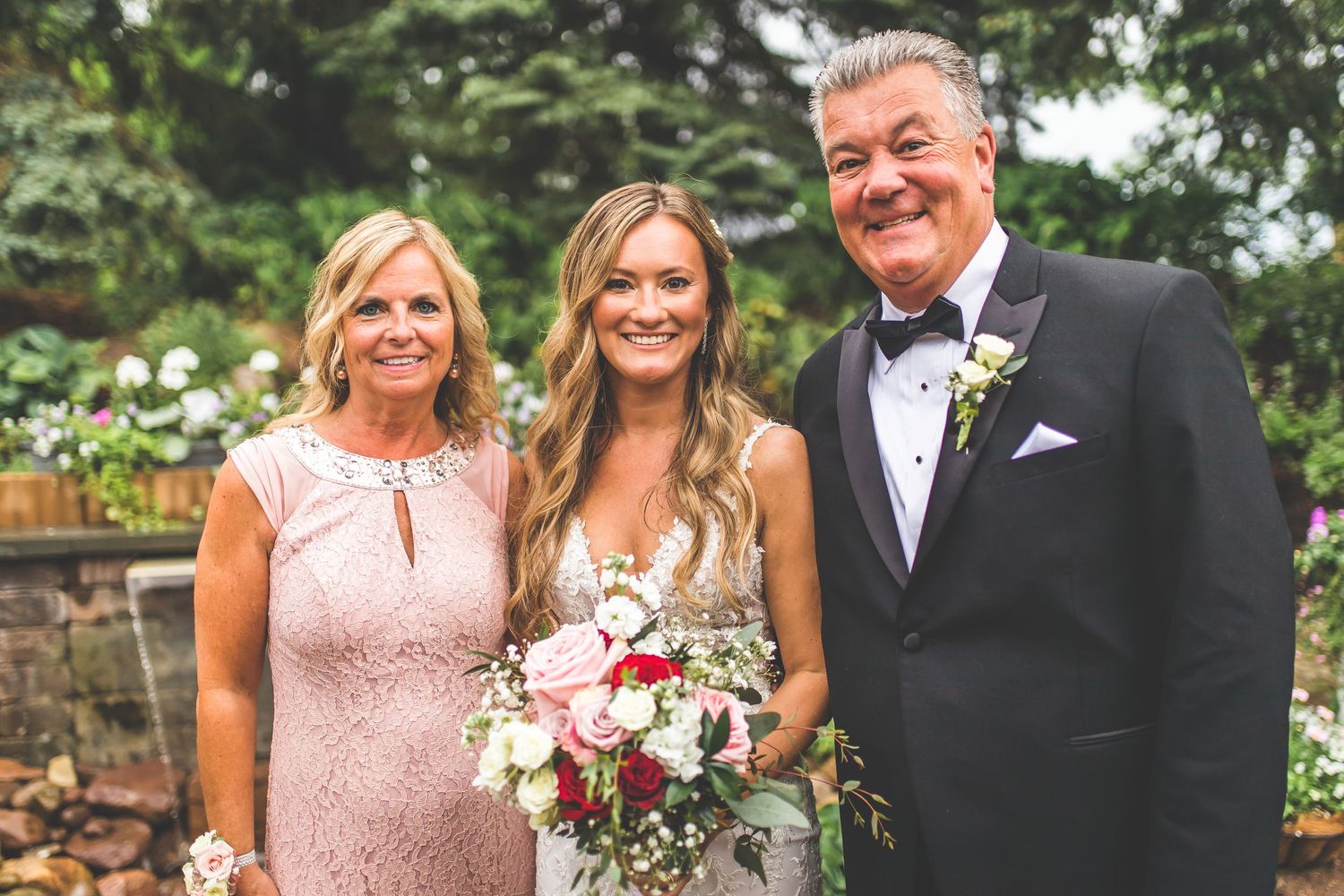 Elegant wedding party photo featuring a bride in white with pink and white bouquet alongside formal-dressed guests outdoors.
