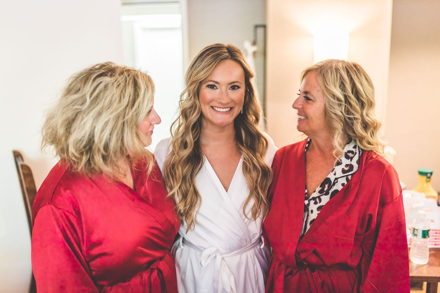 Bridesmaids wearing pink and red robes laugh together during wedding day preparations.