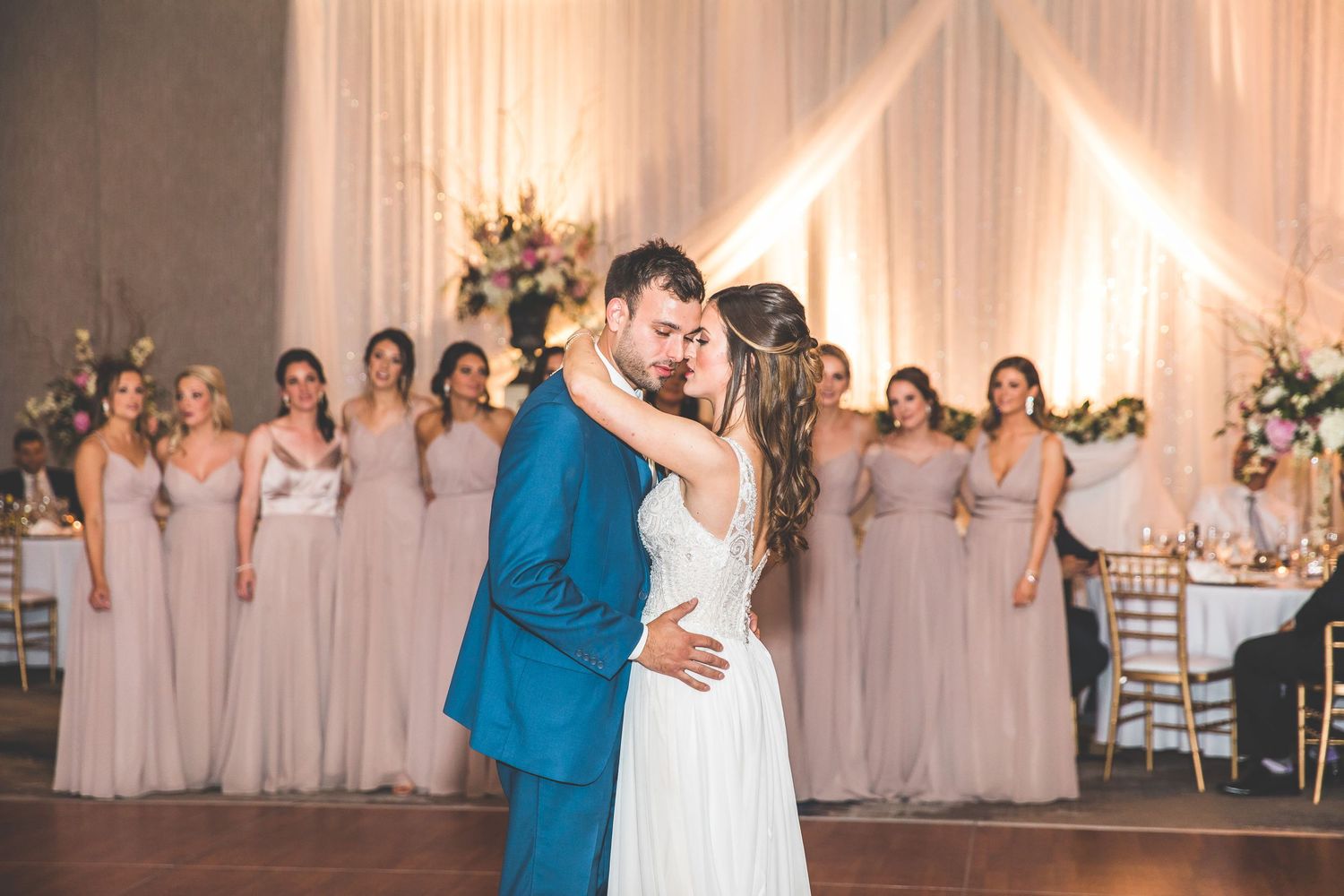 Couple shares first dance surrounded by bridesmaids in blush dresses at elegant reception venue.