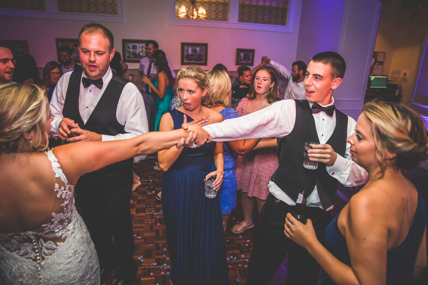 Wedding party members reaching out to join hands while dancing at an indoor reception.