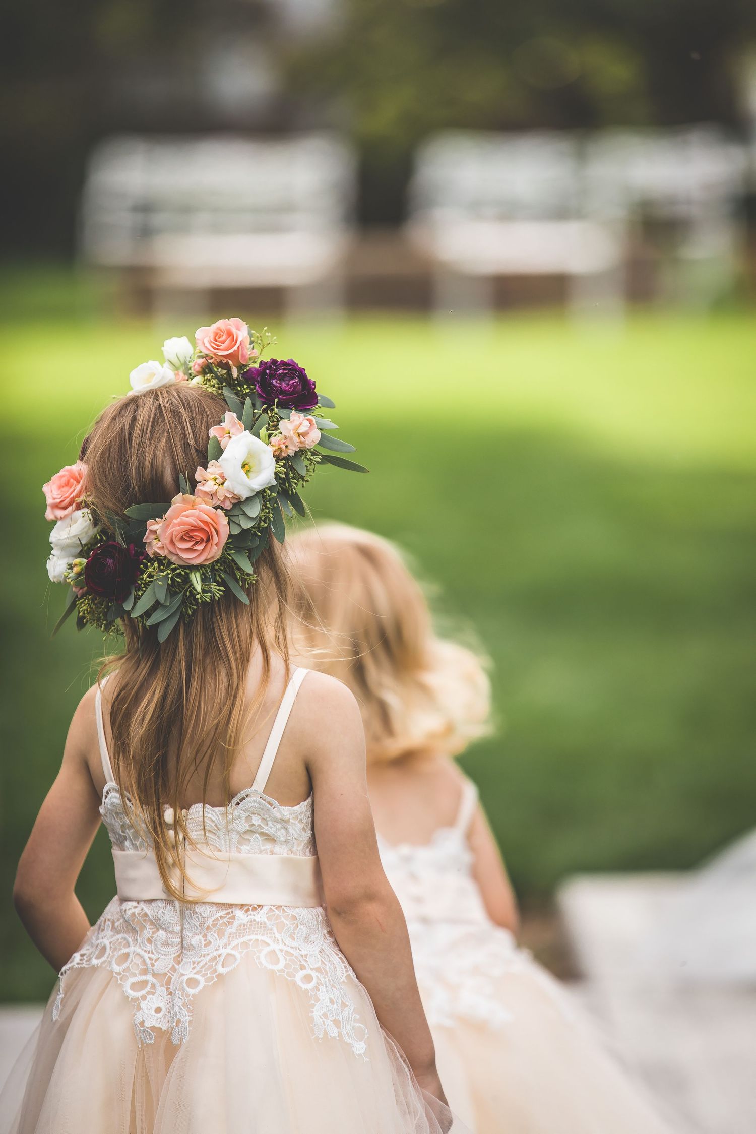 Young flower girls in white dresses wearing floral crowns walk together outdoors.
