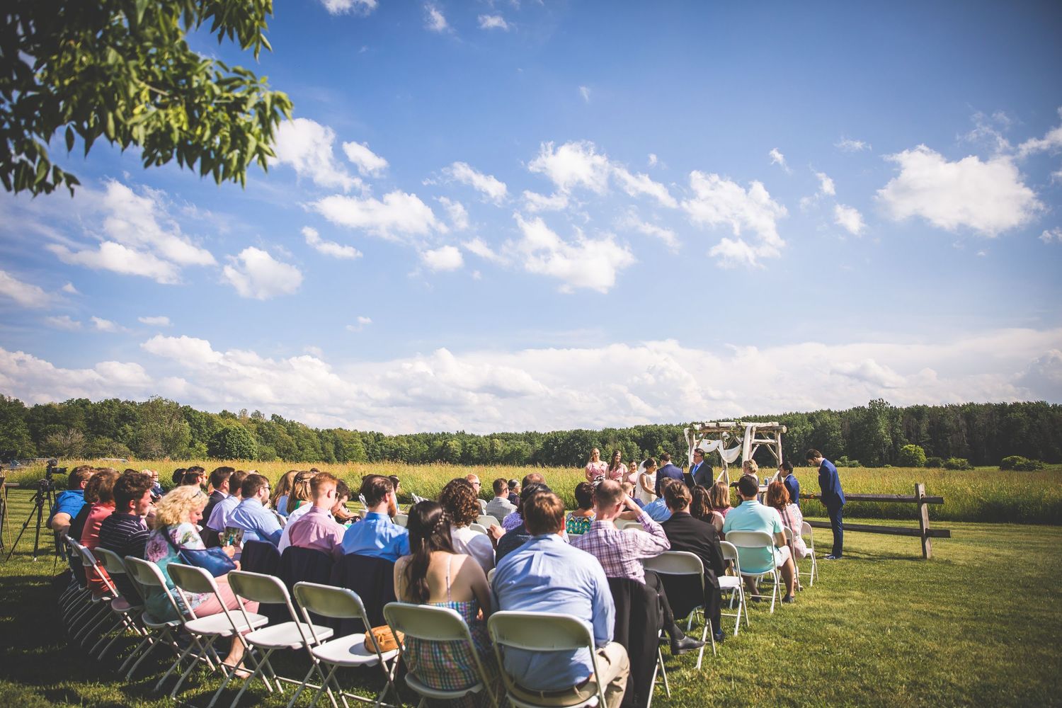 Panoramic view of an outdoor wedding ceremony setup with white chairs and guests on a grassy field.