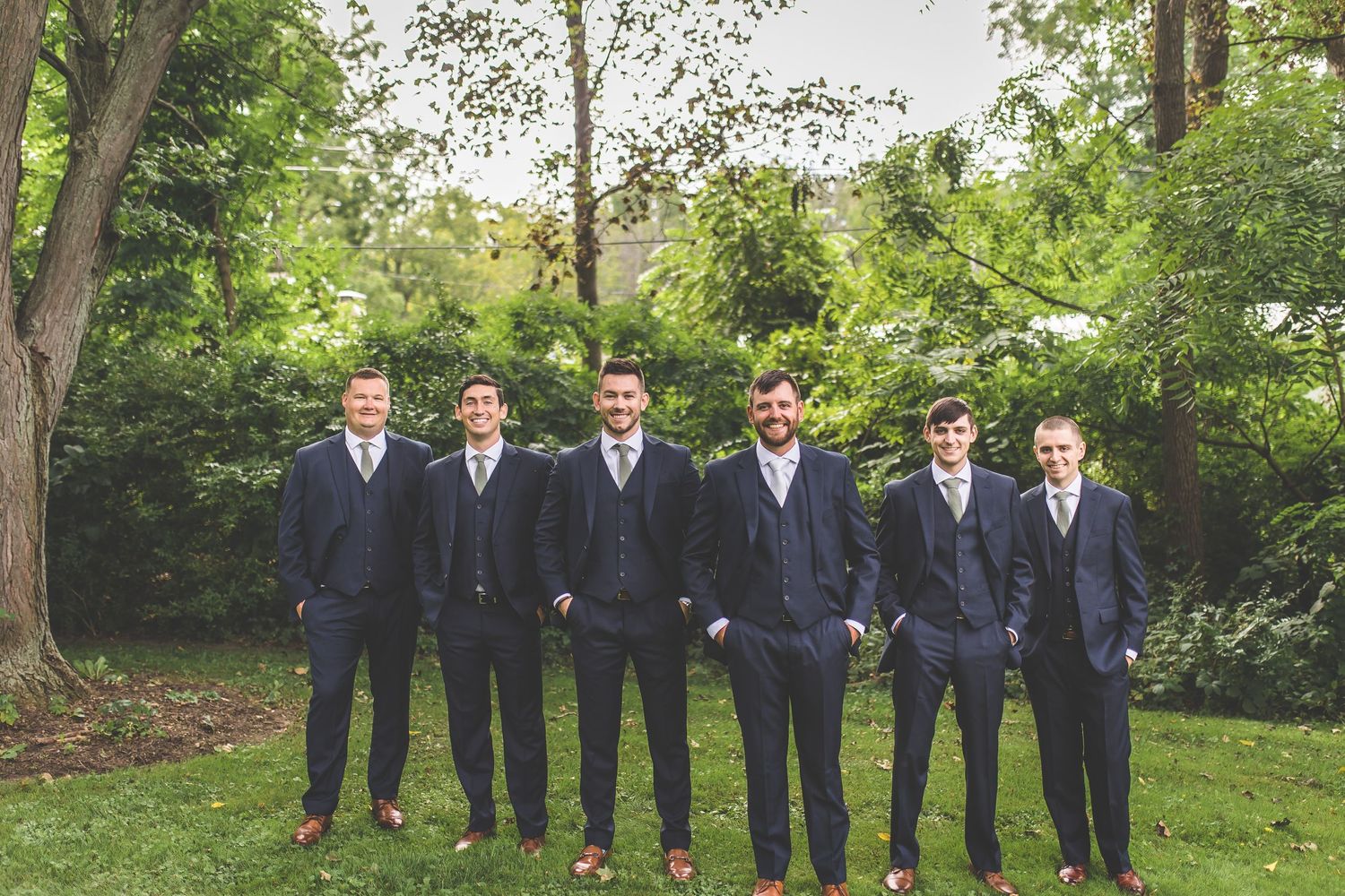 Groomsmen in navy suits pose together outdoors in a garden setting.