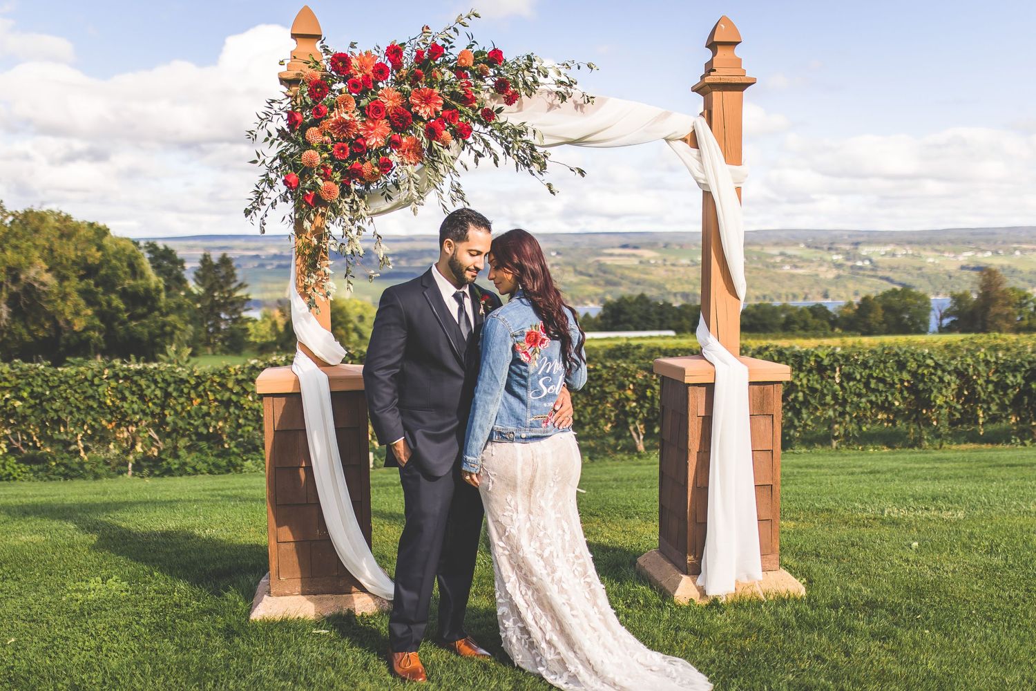 Outdoor wedding ceremony setup with wooden arch decorated with vibrant red flowers against scenic landscape.