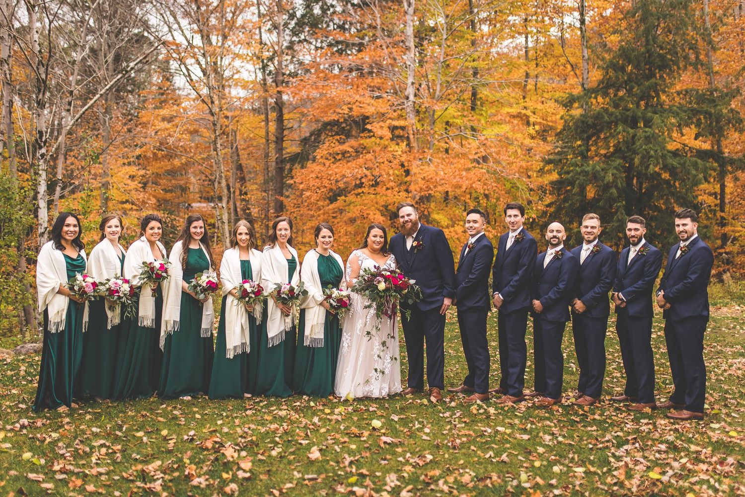 Wedding party poses in front of vibrant fall foliage wearing navy and emerald attire.