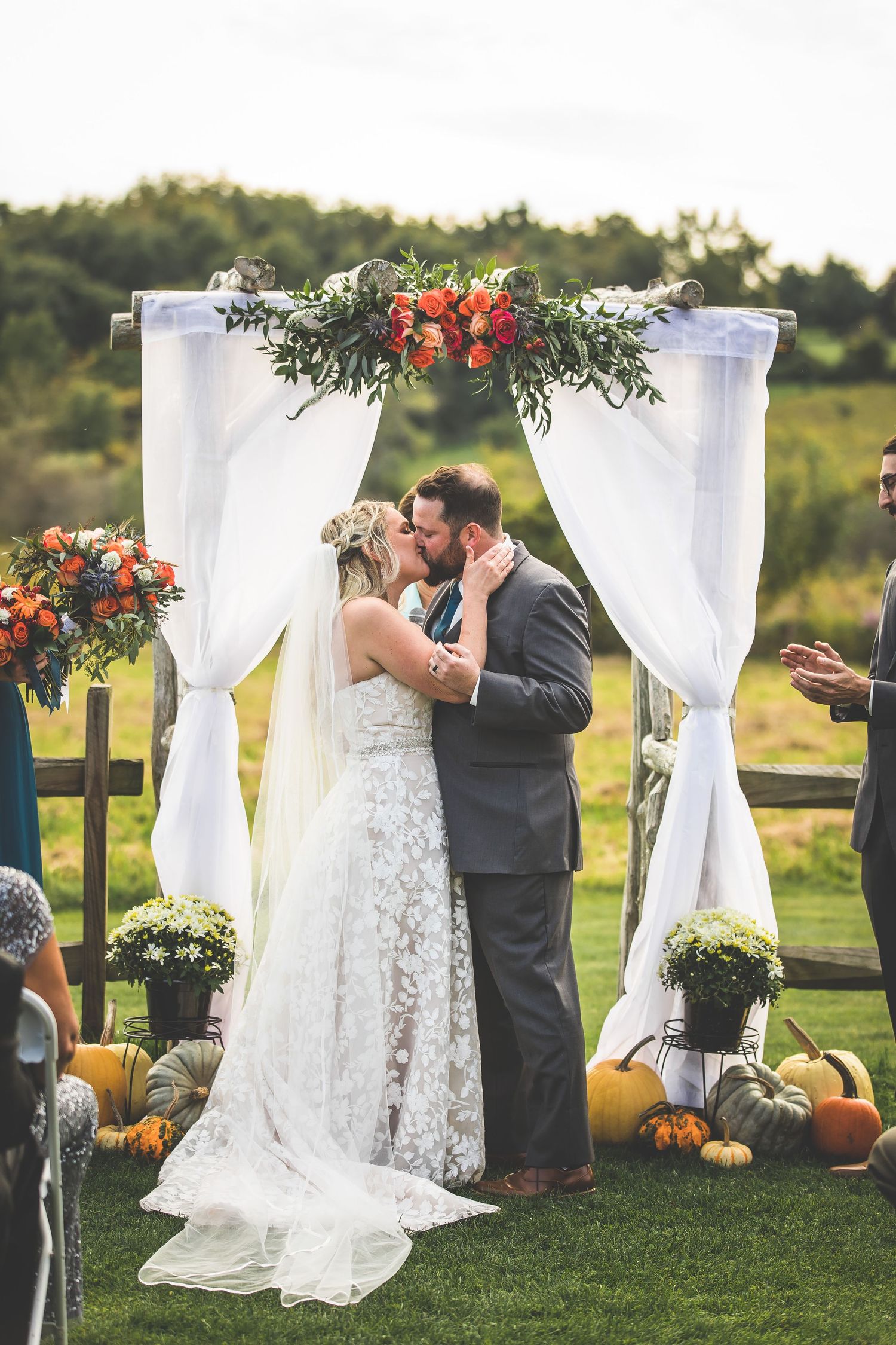 Wedding ceremony site decorated with white drapes, pumpkins, and orange floral arrangements.