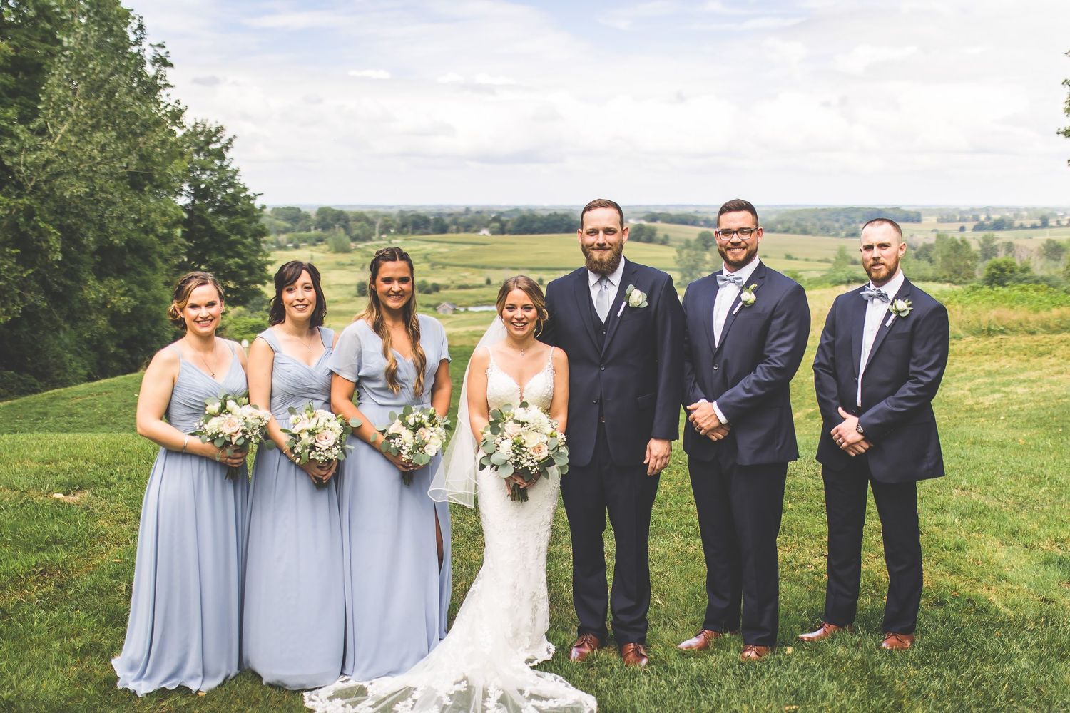 Wedding party in light blue bridesmaid dresses and navy suits poses together on a grassy hillside.