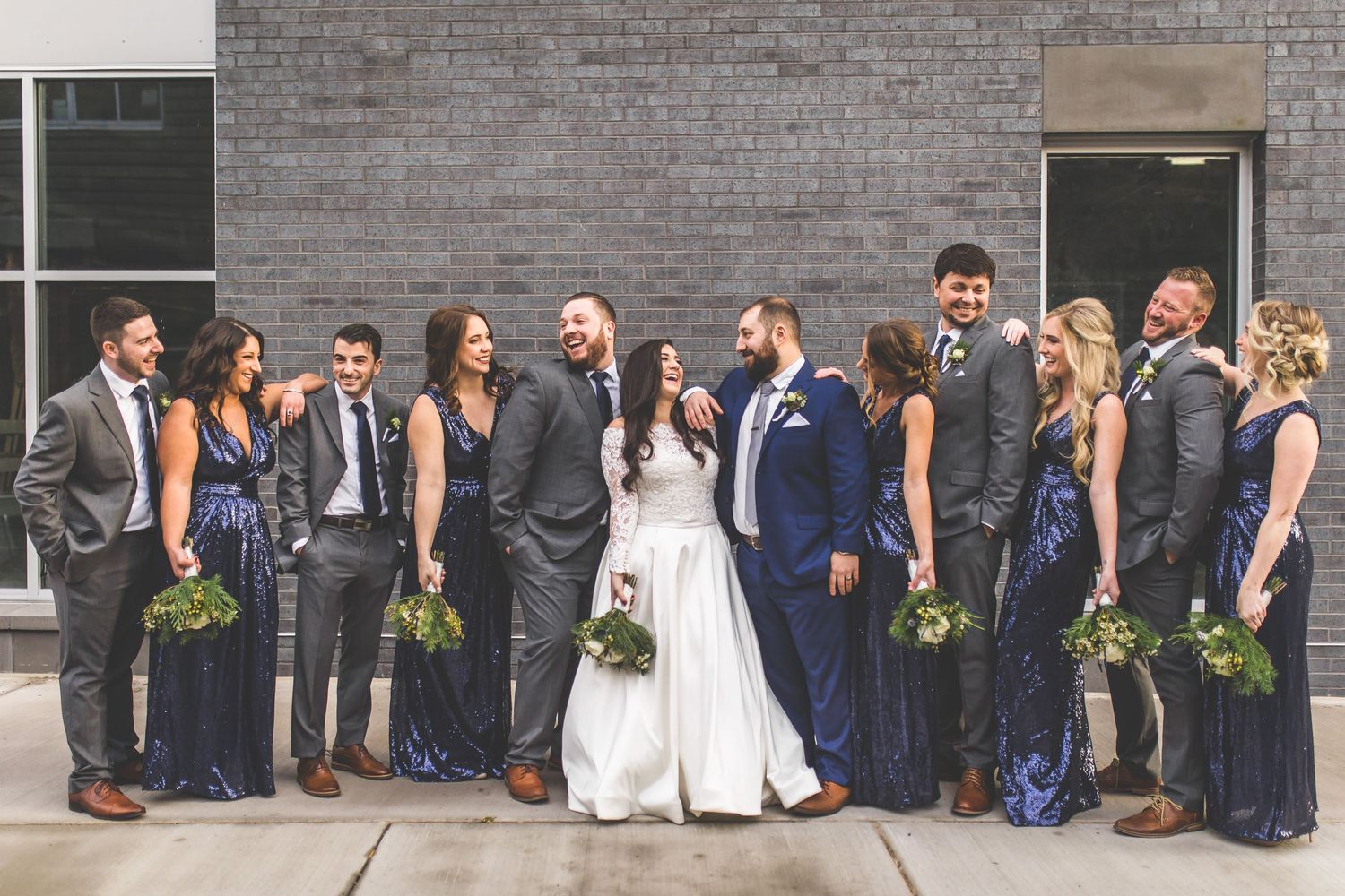 Stylish wedding party in navy and gray attire poses casually against modern brick building.