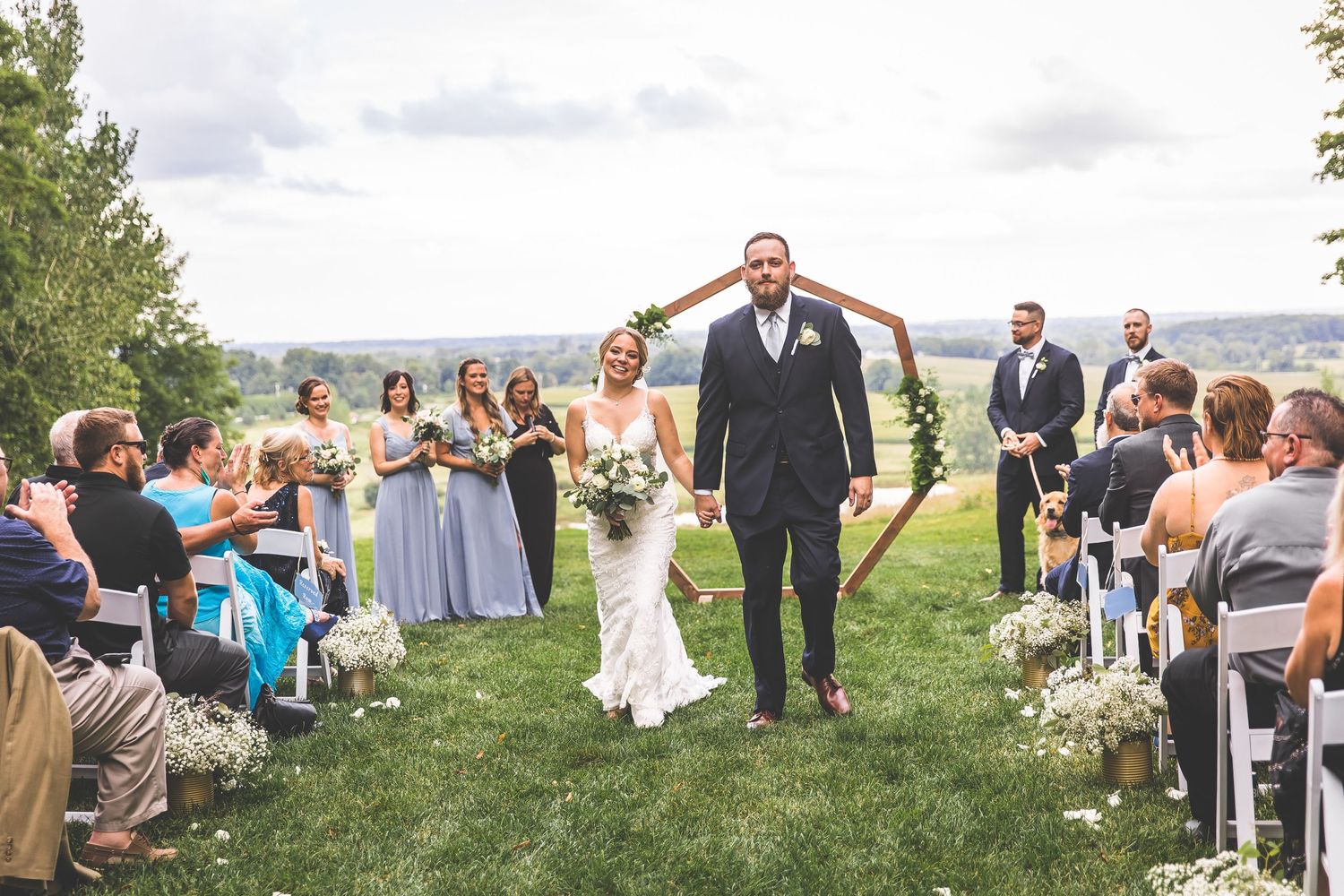 Wedding celebration with guests seated outdoors as newlyweds celebrate under wooden arch.