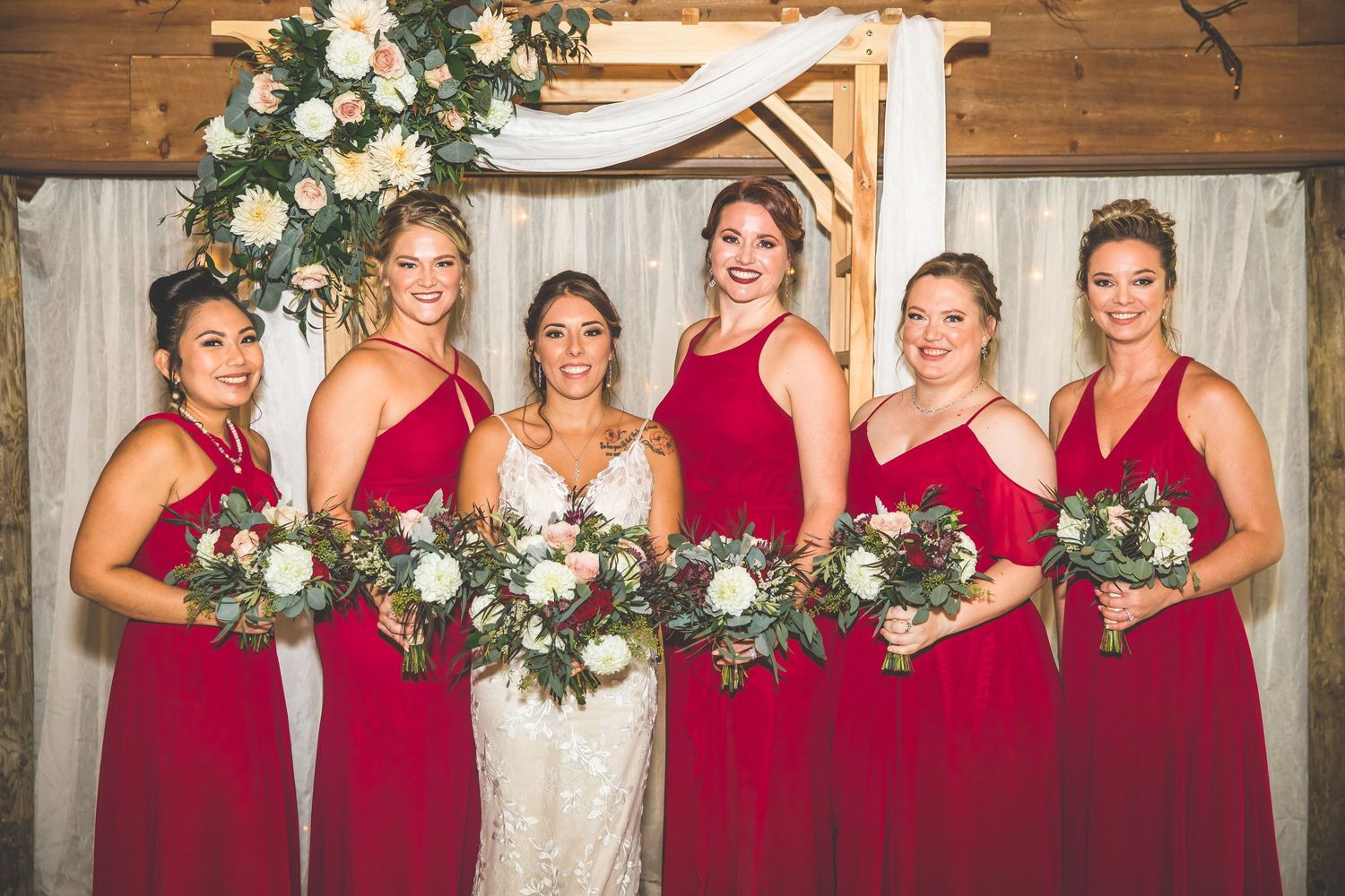Bridesmaids in matching red gowns pose with bride in rustic wooden venue with floral decorations.
