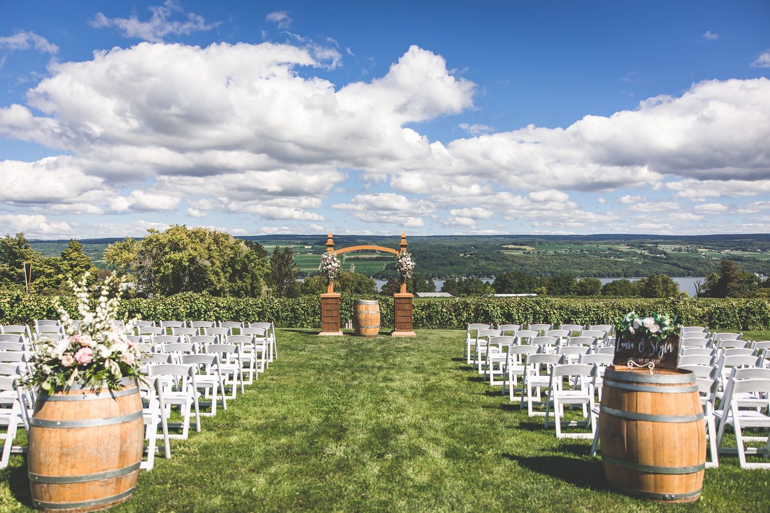Outdoor wedding ceremony setup with white chairs and wine barrels under blue sky.