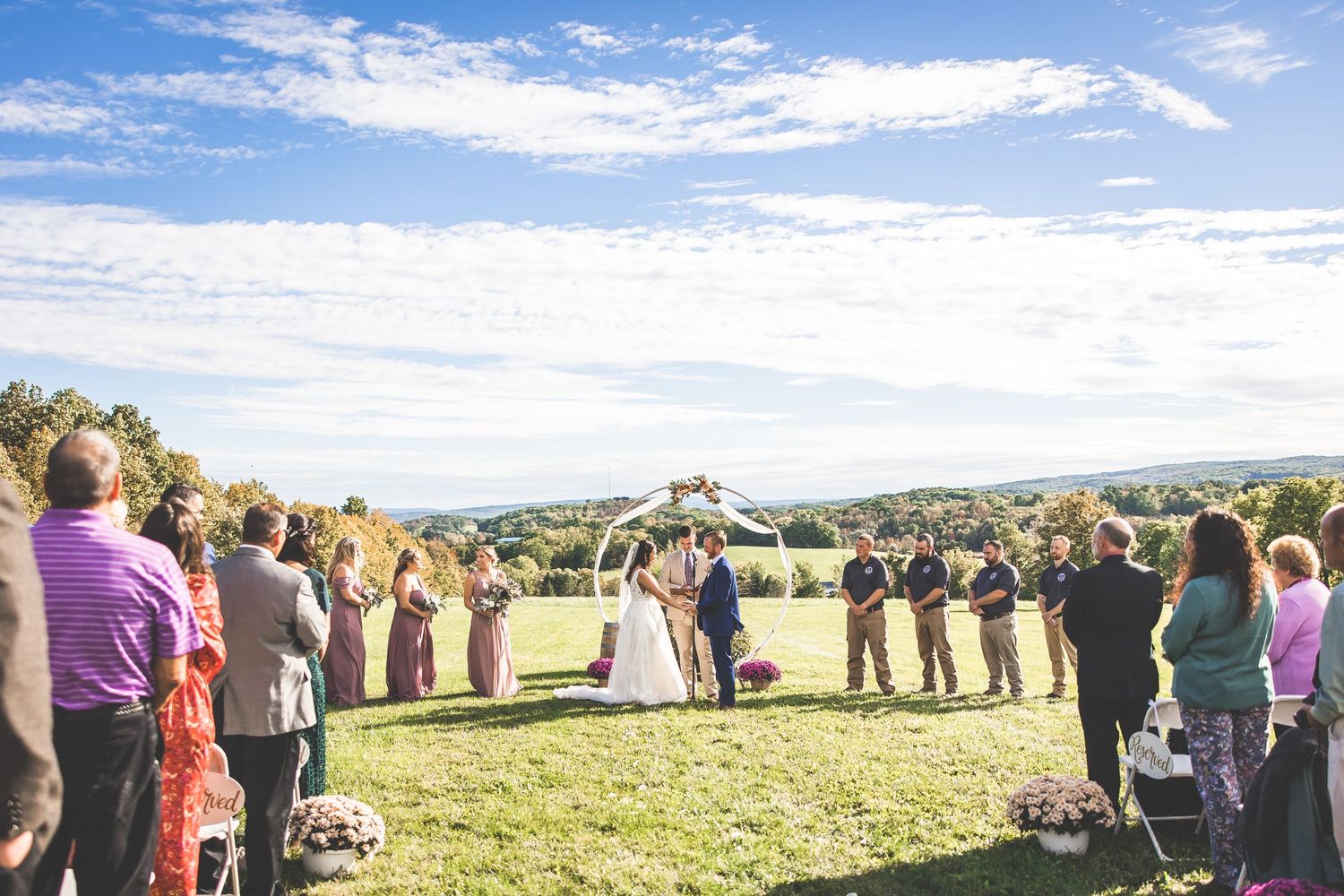 Panoramic view of a wedding ceremony on a grassy hillside with guests surrounding the couple.