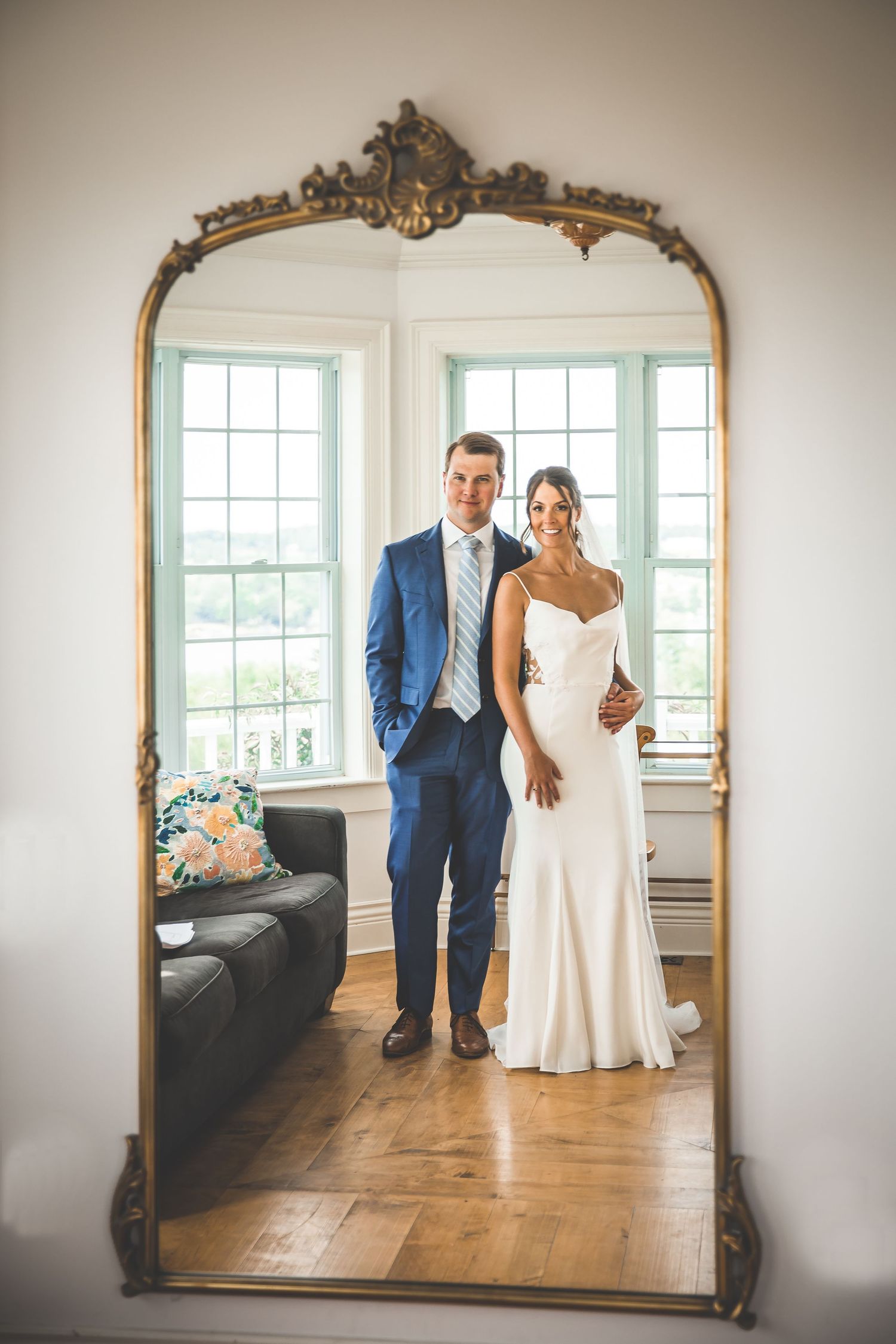Elegant ornate mirror reflects a couple in a bright white room with hardwood floors.