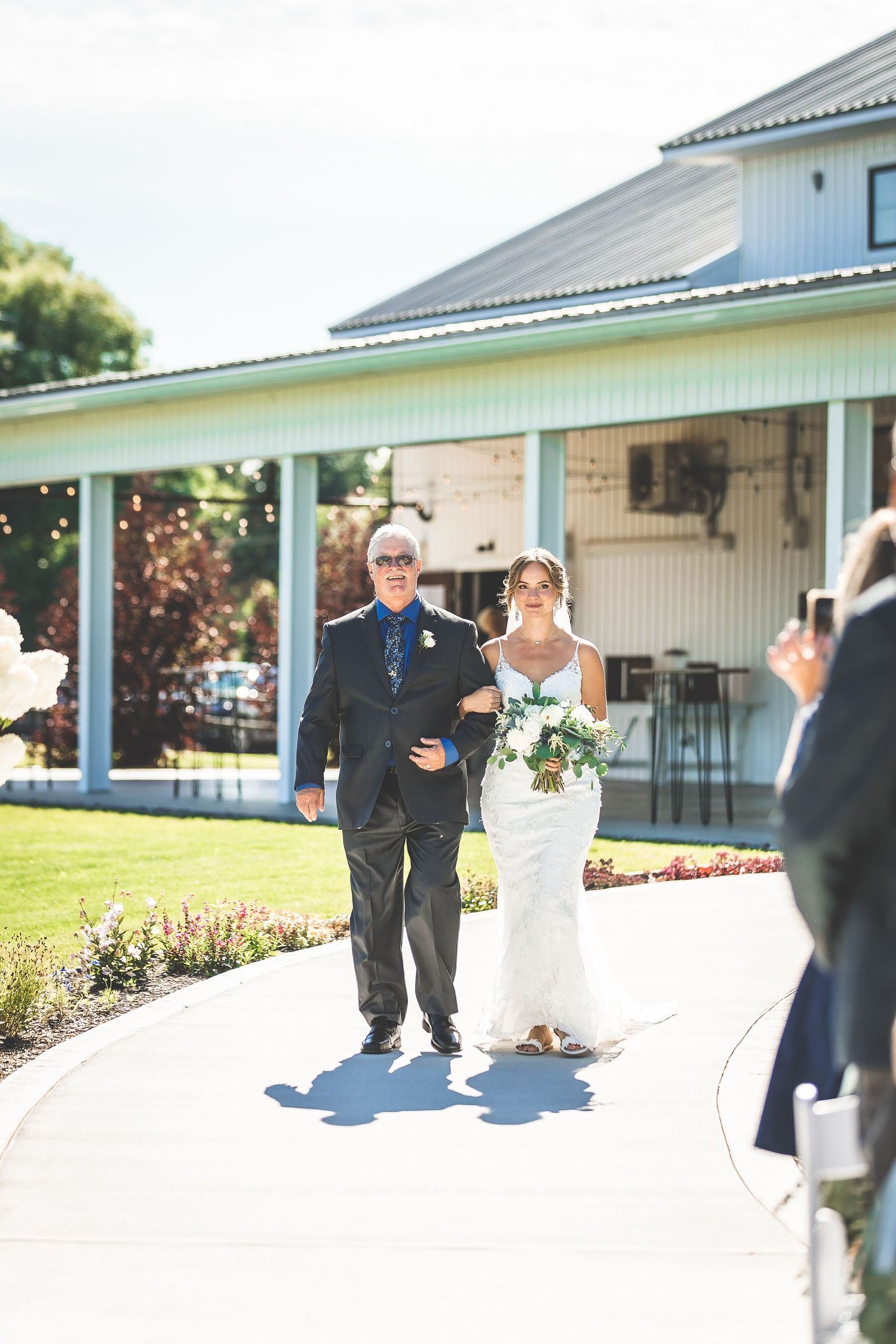 Outdoor wedding ceremony entrance with couple walking down path near blue and white building.