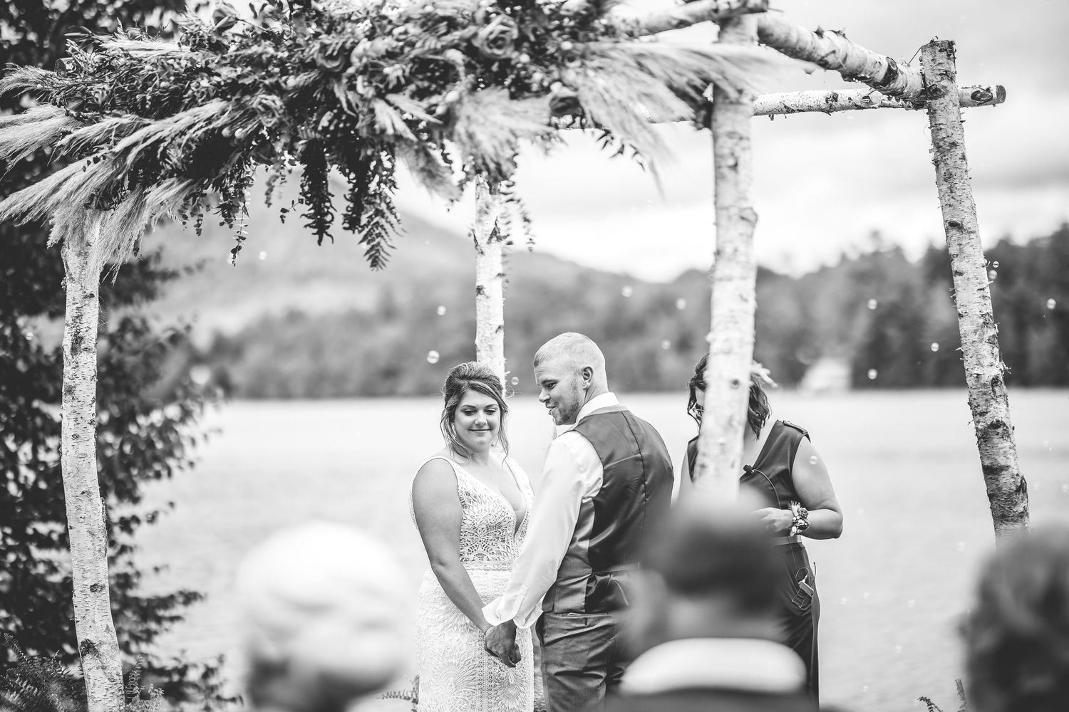 Black and white photo of a wedding ceremony taking place under a decorated birch arch by a serene lake.