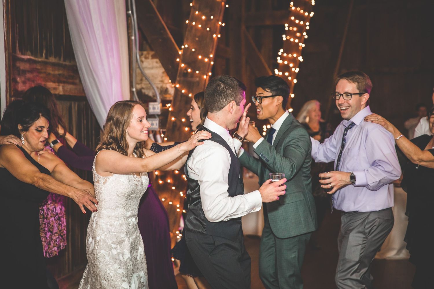 Wedding guests dance and celebrate in a rustic venue decorated with string lights.