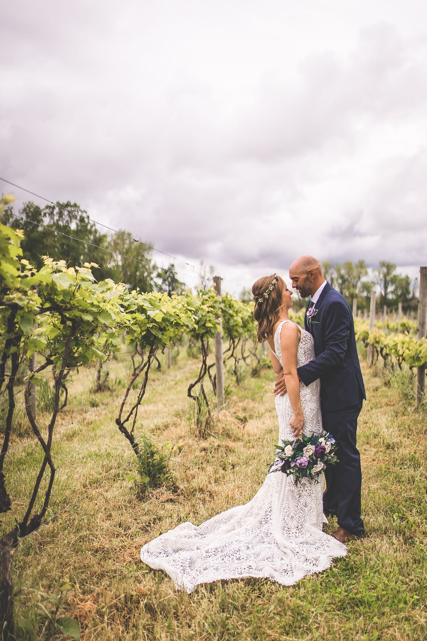 Romantic wedding couple embraces among grapevines on an overcast day in a vineyard.