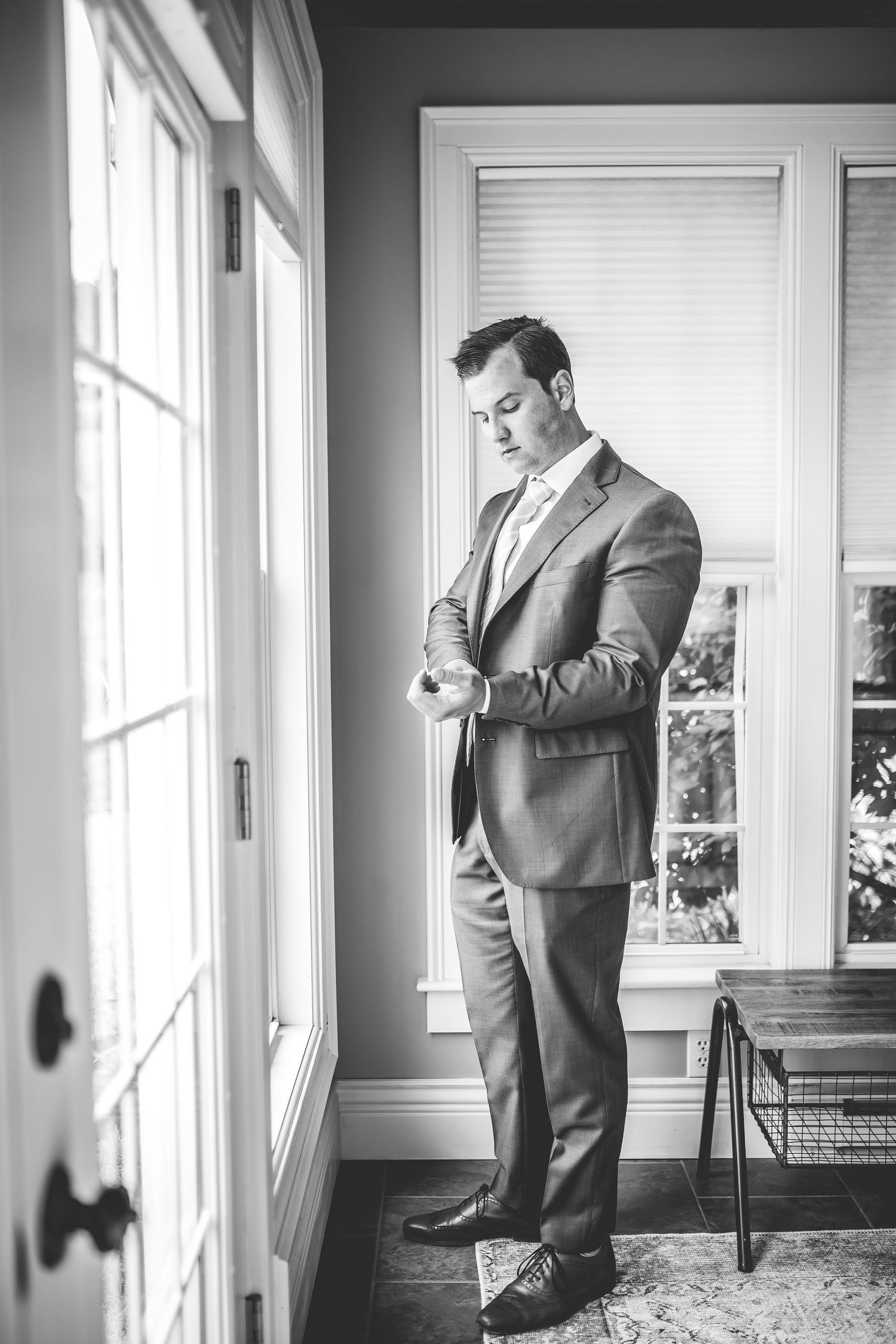 Groom in classic grey suit adjusts his tie while standing in a sunlit room.