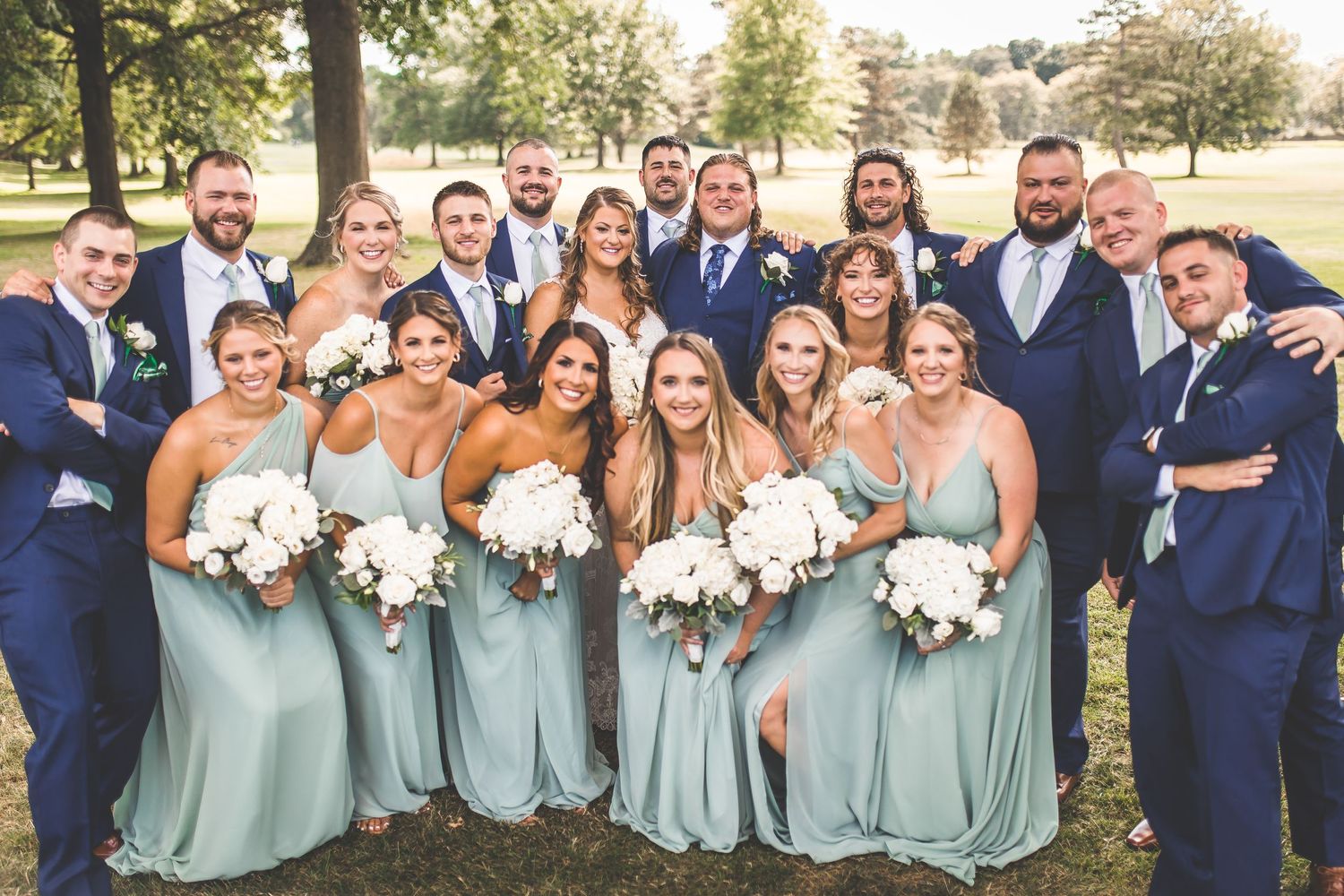 Large wedding party in light blue dresses and navy suits gathered for a group photo outdoors.