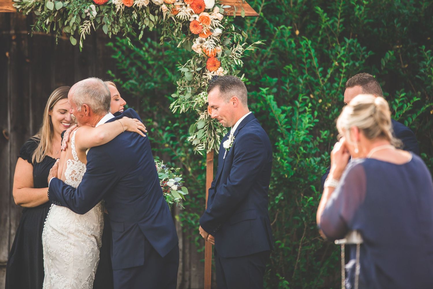 Wedding ceremony under a wooden arch decorated with coral and white floral arrangements.