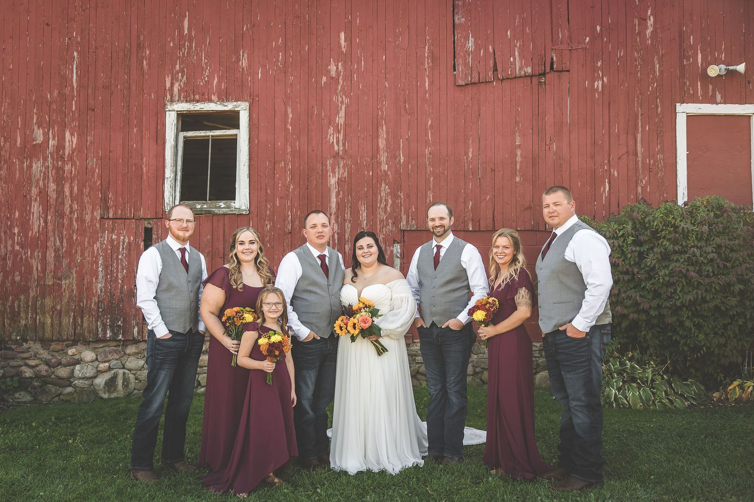 Wedding party in burgundy and gray attire poses in front of rustic red barn.
