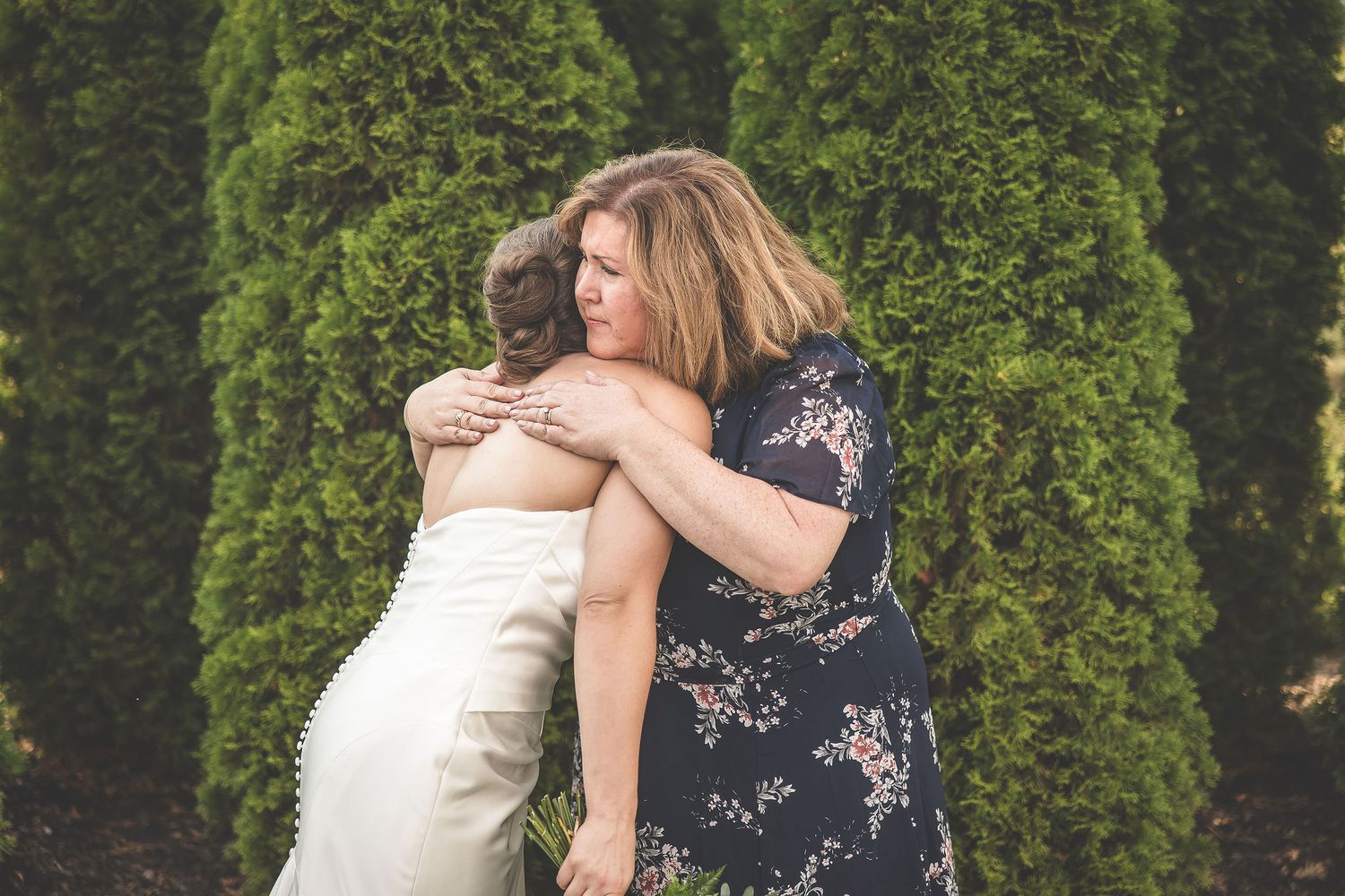 Emotional embraces shared between wedding party members in front of green hedges.