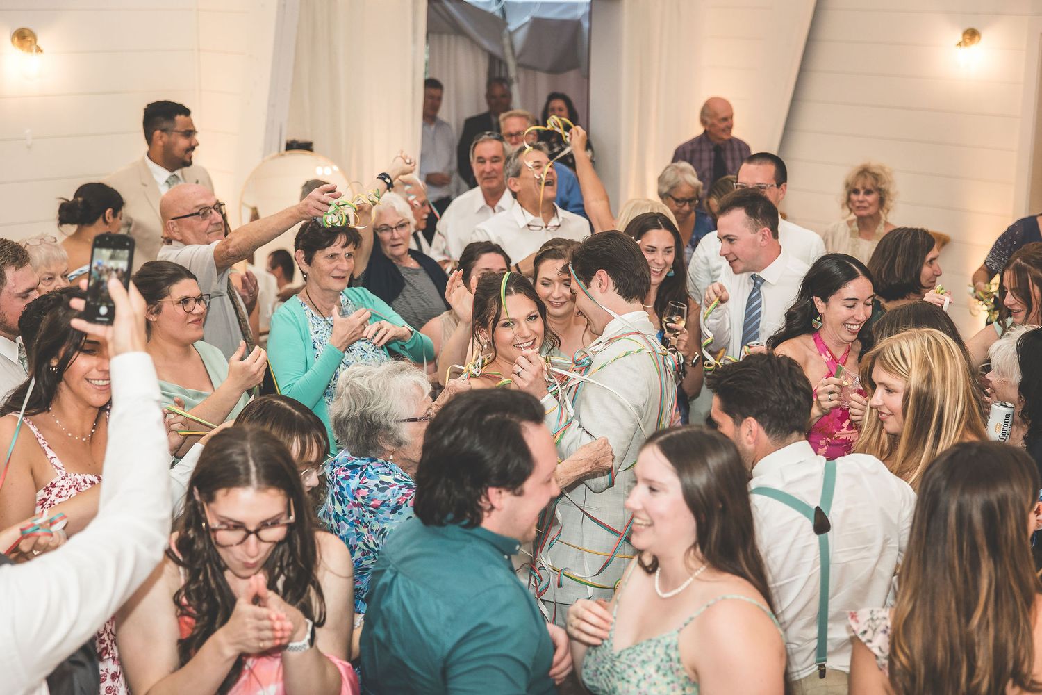 Large group of wedding guests dancing and mingling at an indoor reception with white walls.