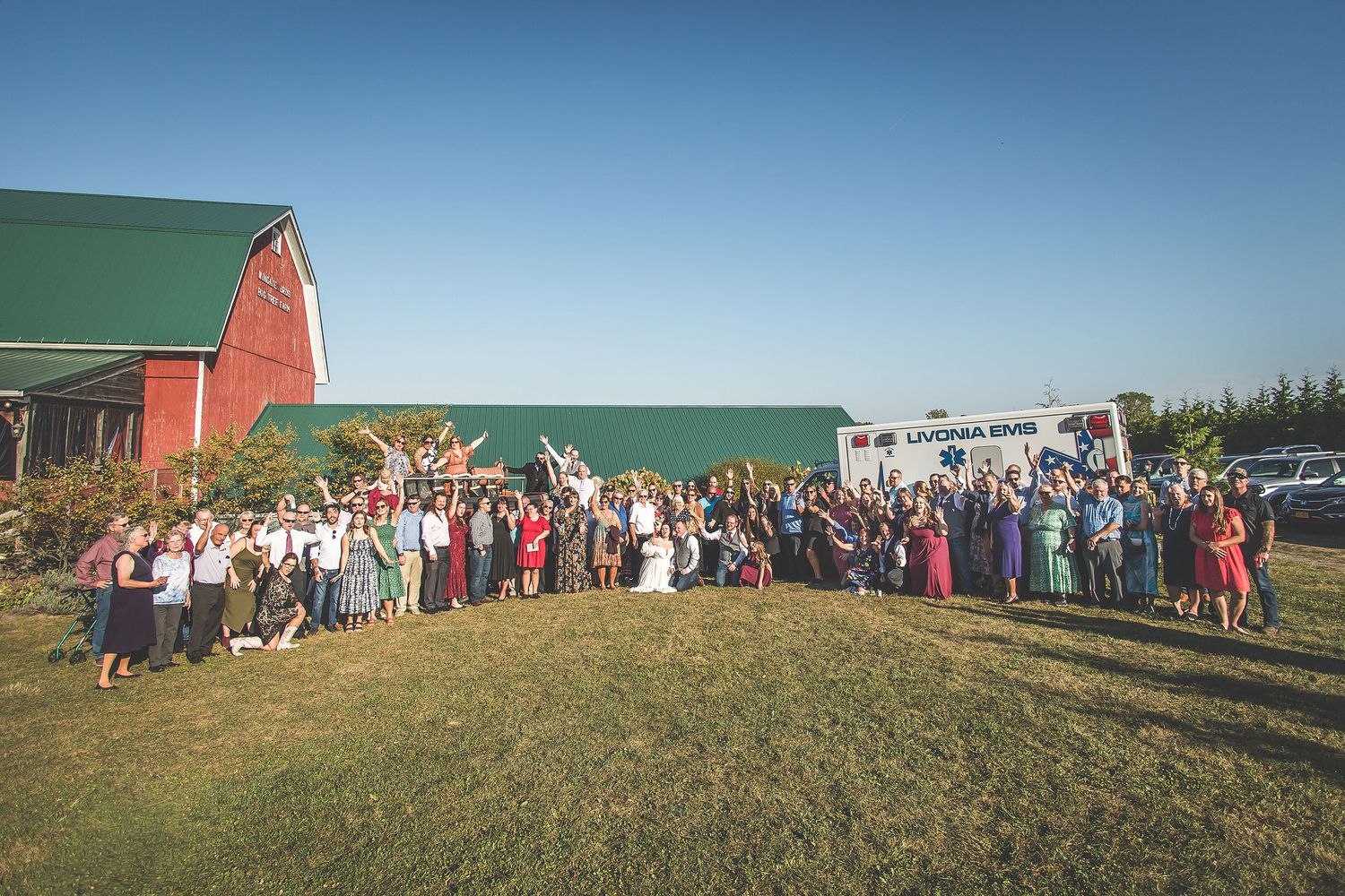 Large group photo of wedding guests and first responders lined up on a grassy hill by a barn.