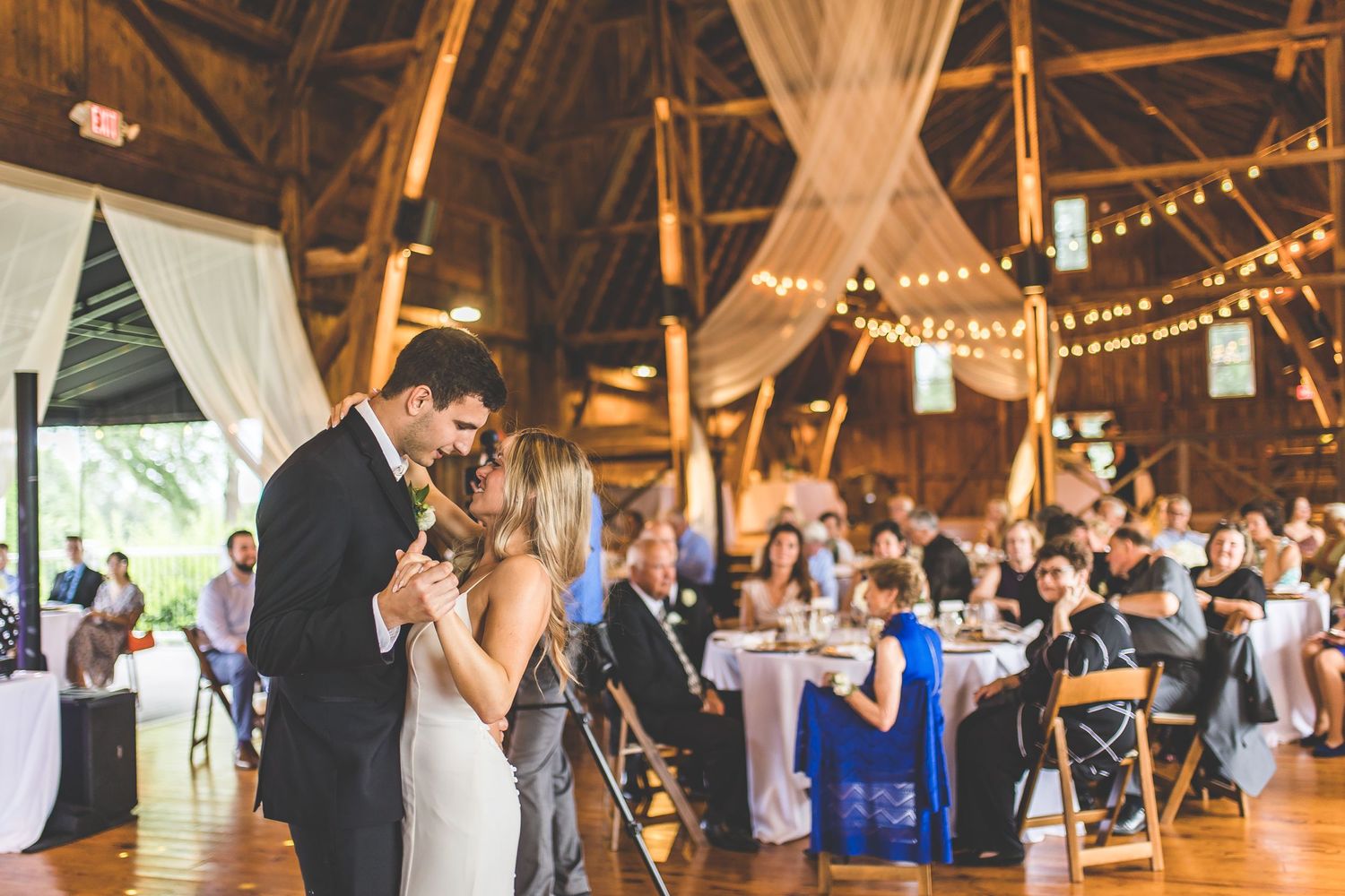 Couple dancing together during wedding reception in rustic barn venue with string lights and white decor.