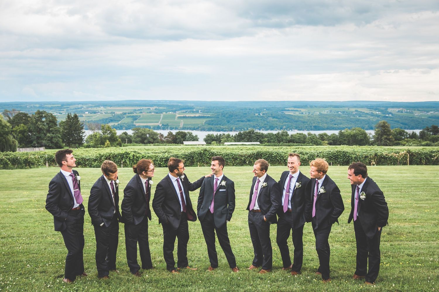 Groomsmen in dark suits standing on hilltop with scenic lake view in background.
